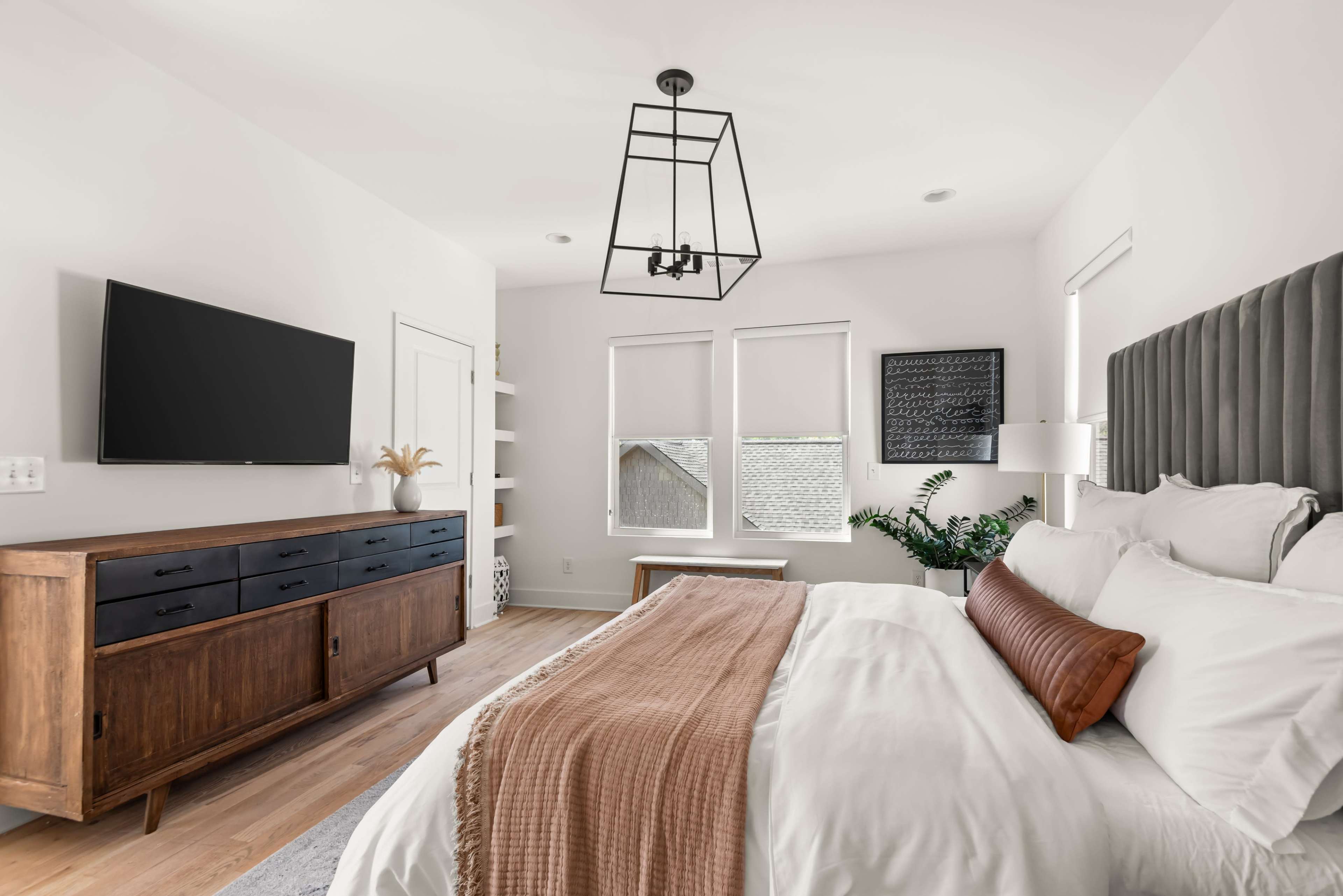 The image shows a modern bedroom featuring a large bed with white linens, a wooden dresser with a TV mounted above it, and natural light coming through the windows.