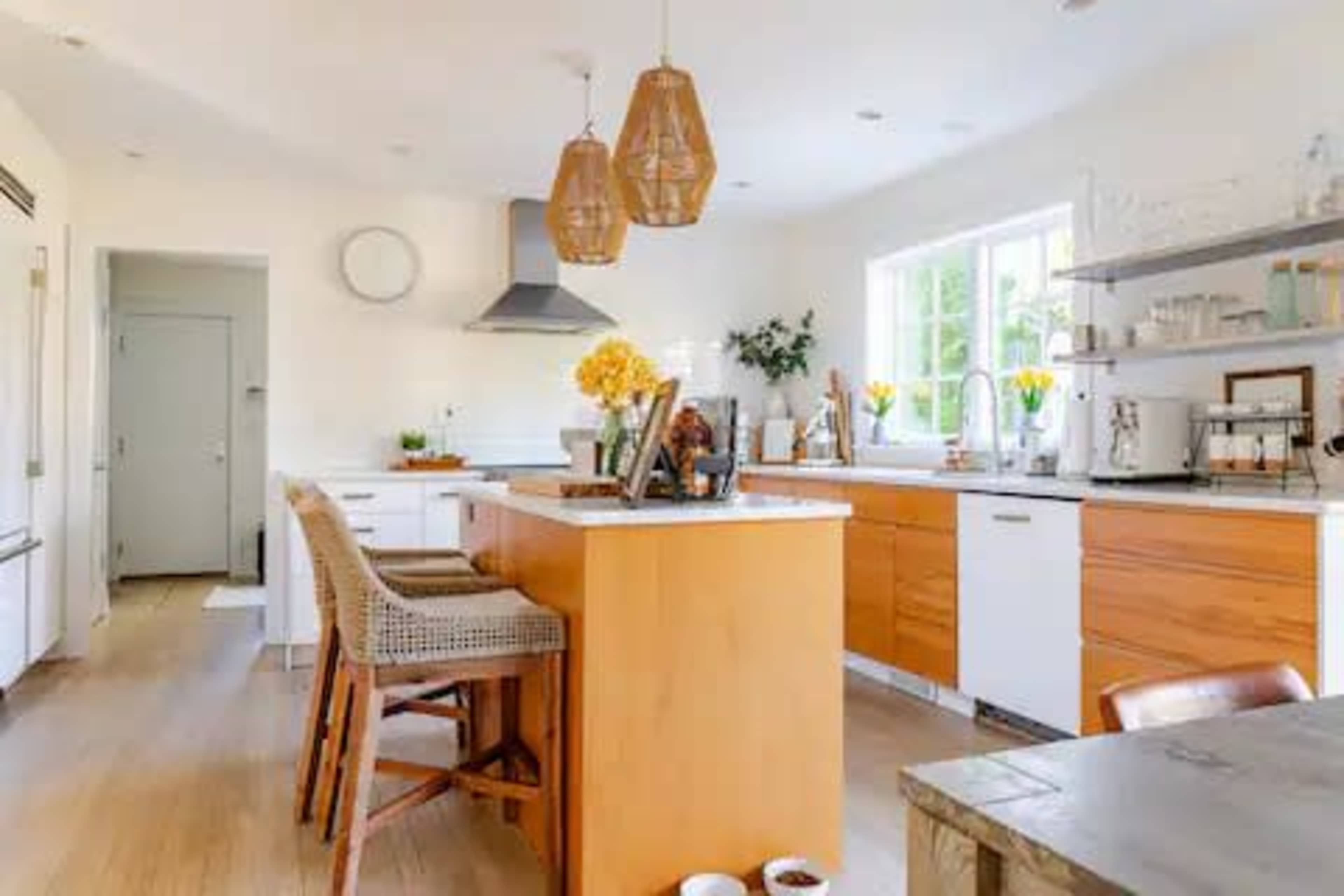 The kitchen features a central island with wooden cabinetry, hanging pendant lights, and modern appliances, surrounded by light-colored walls and a large window.