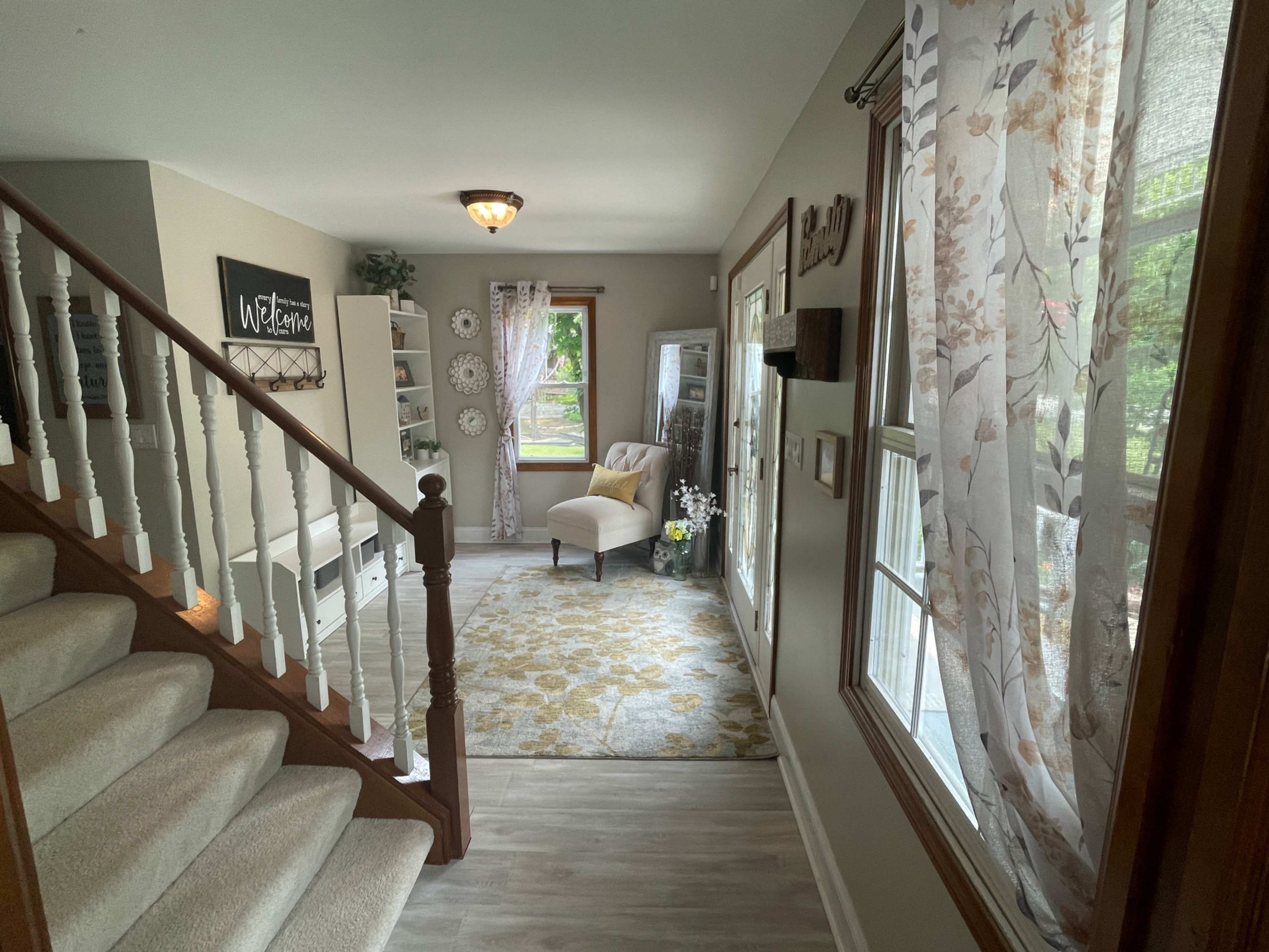 The image shows a bright entryway with a staircase, a cozy chair, and large windows draped with sheer curtains.