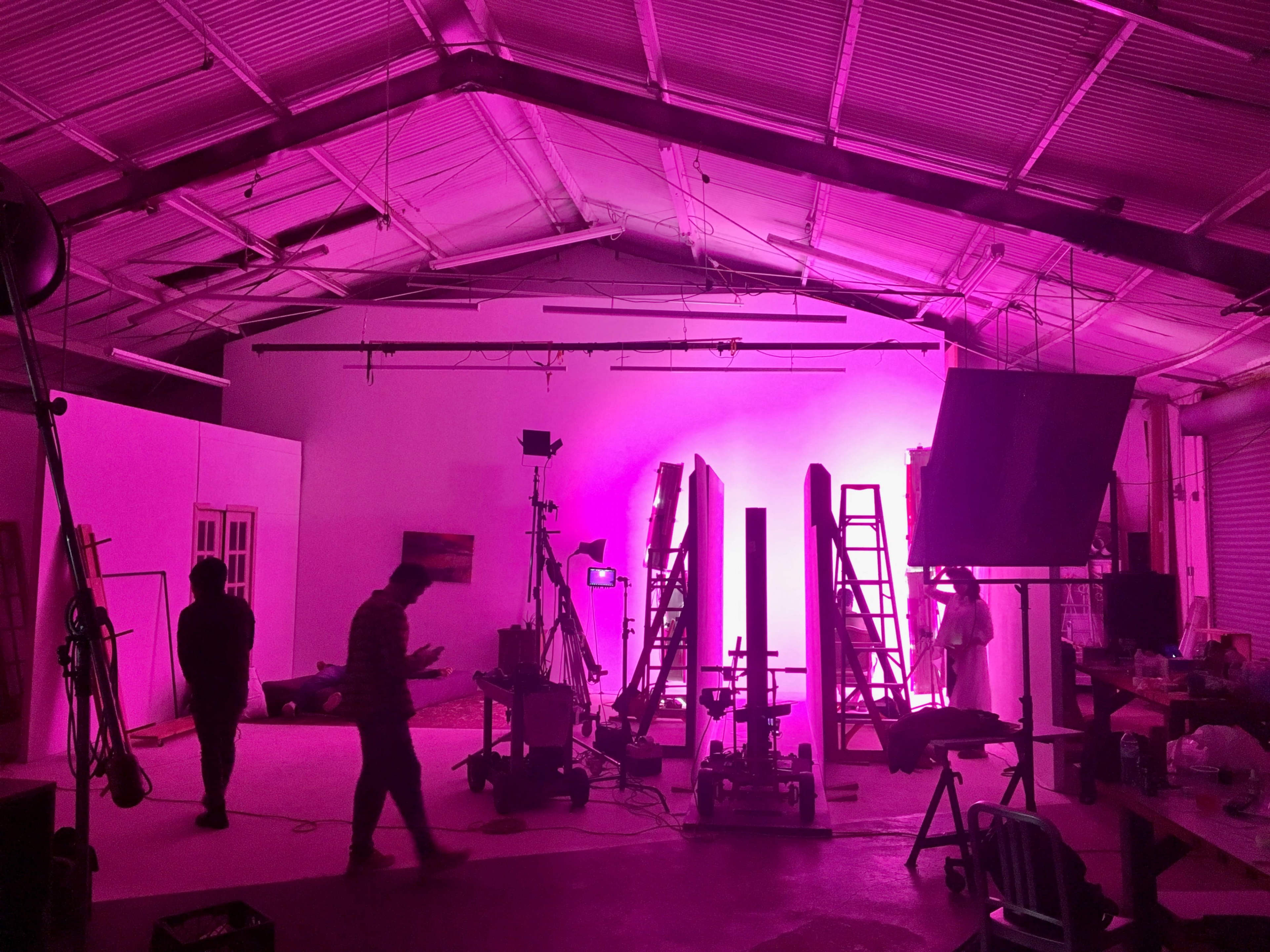 The image shows a dimly lit studio space featuring pink lighting, several ladders, equipment setup, and two people moving around.