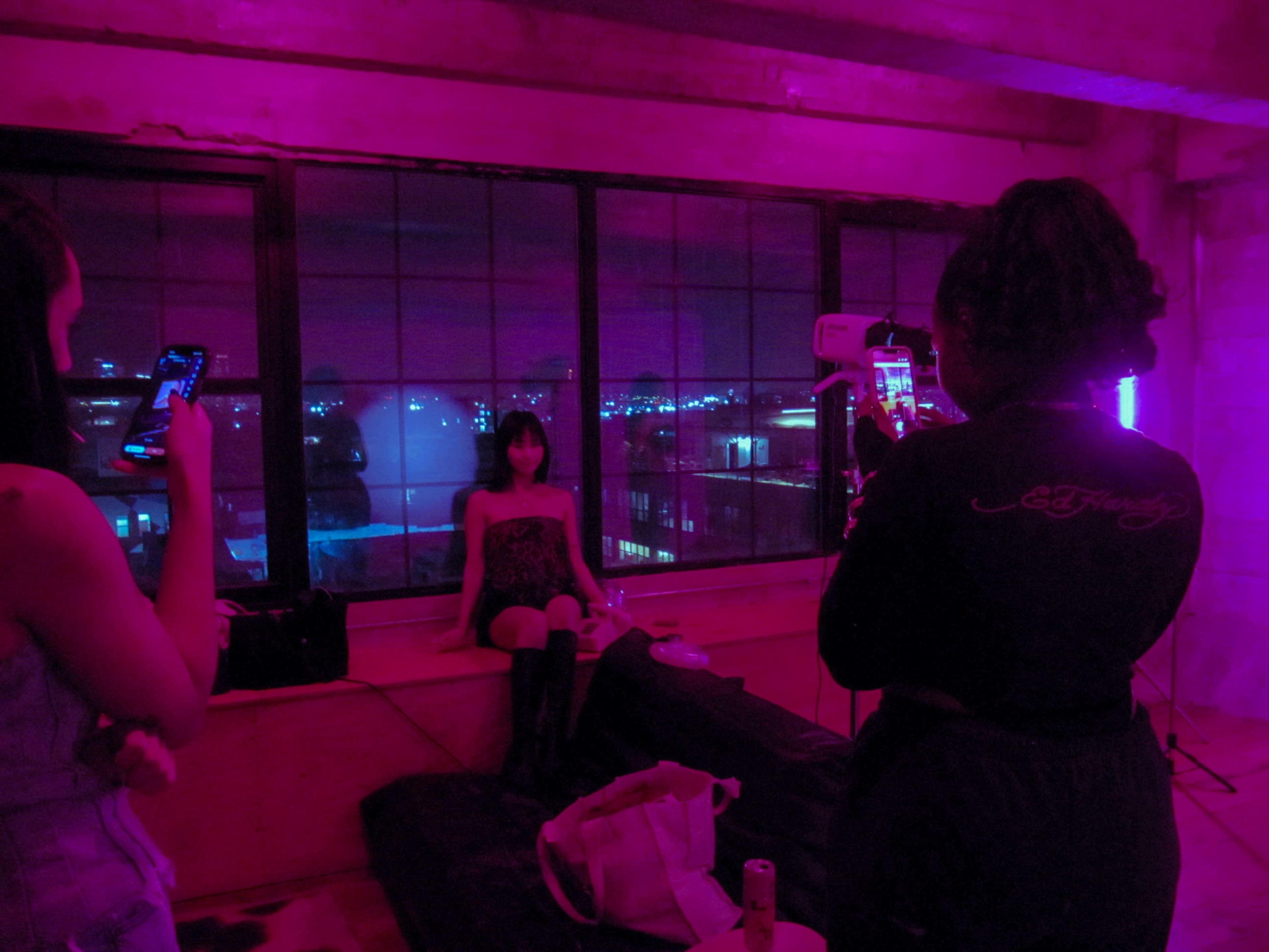 Three people are in a dimly lit room with purple lighting, with one person posing on a bed while the others take photos using their phones.