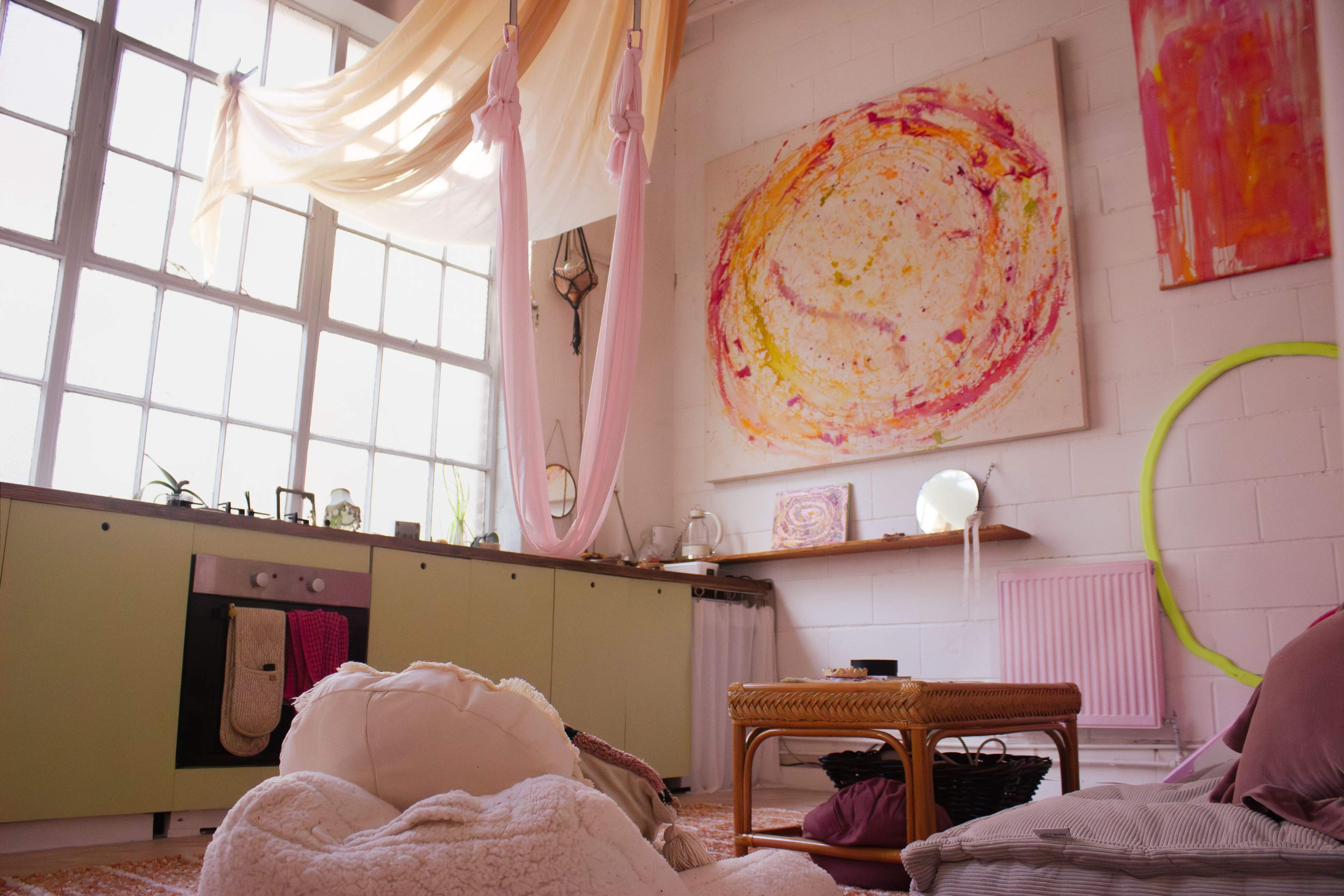 The image shows a cozy, brightly lit room with large windows, featuring a kitchen area, an abstract painting on the wall, and a variety of soft furnishings scattered on the floor.