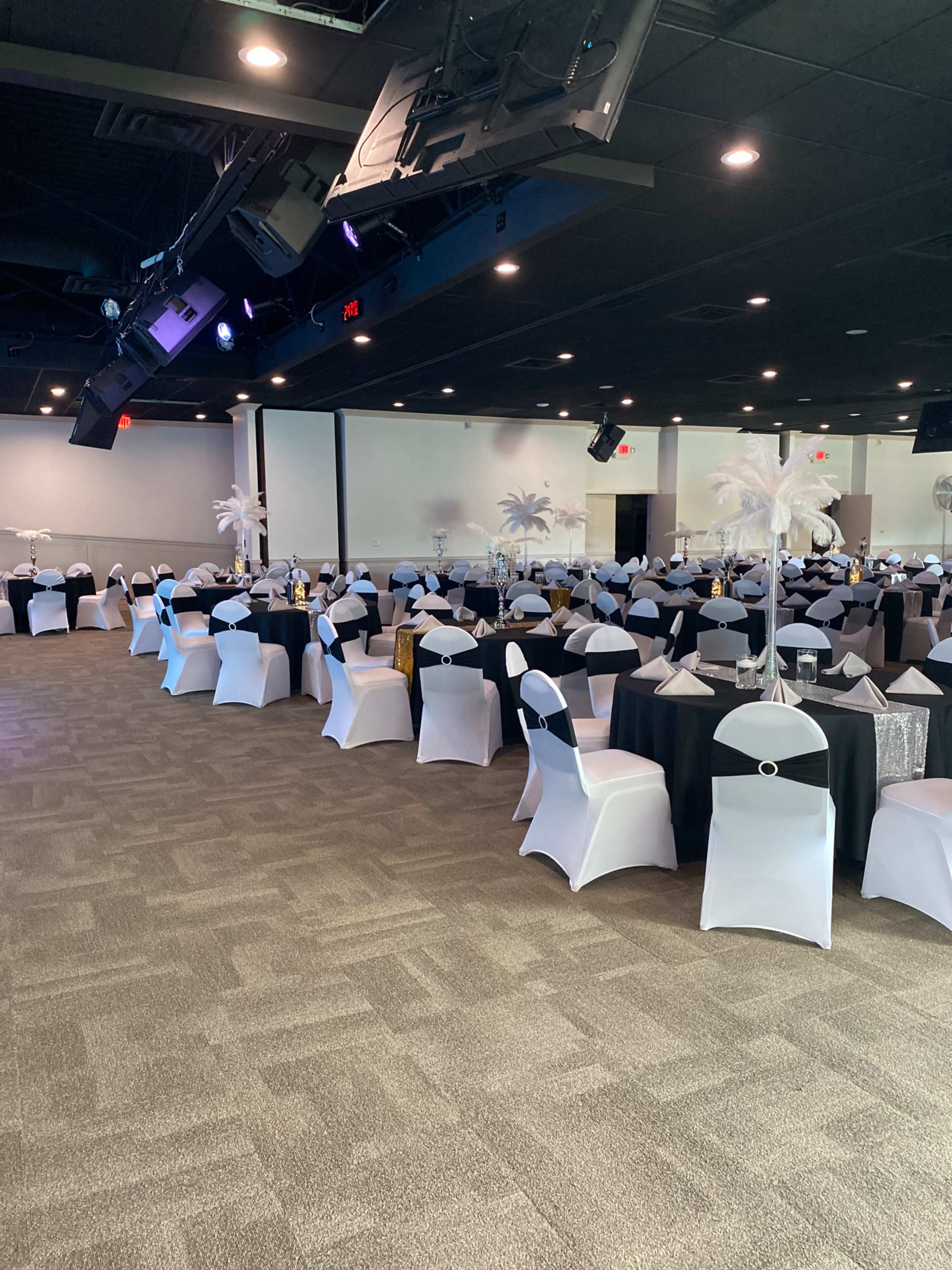 A banquet hall arranged with rows of tables covered in black and white tablecloths, adorned with decorative centerpieces and chairs covered in white and black sashes.