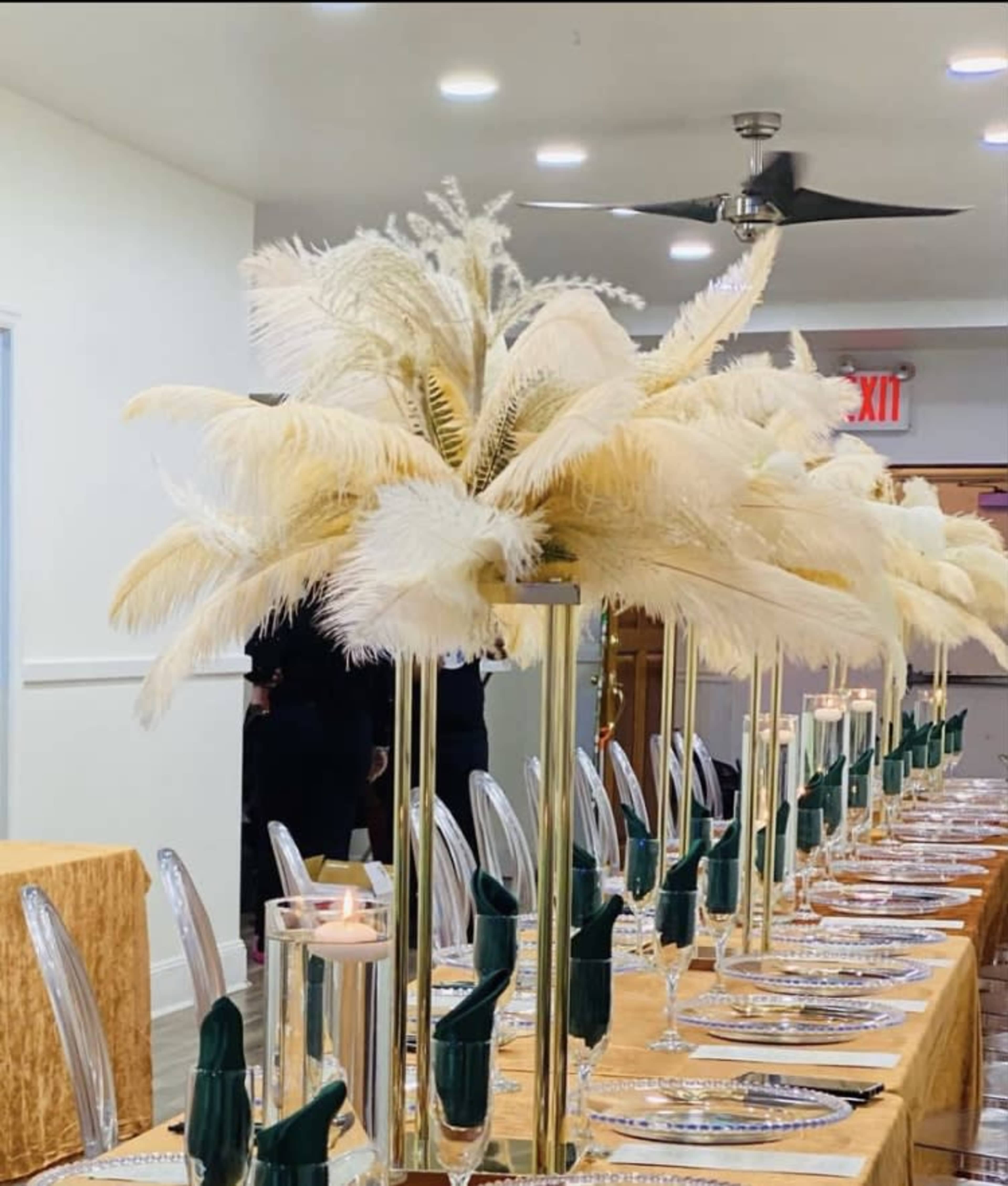 A long dining table is elegantly set with gold tablecloths, transparent plates, and tall centerpieces featuring white feathers.