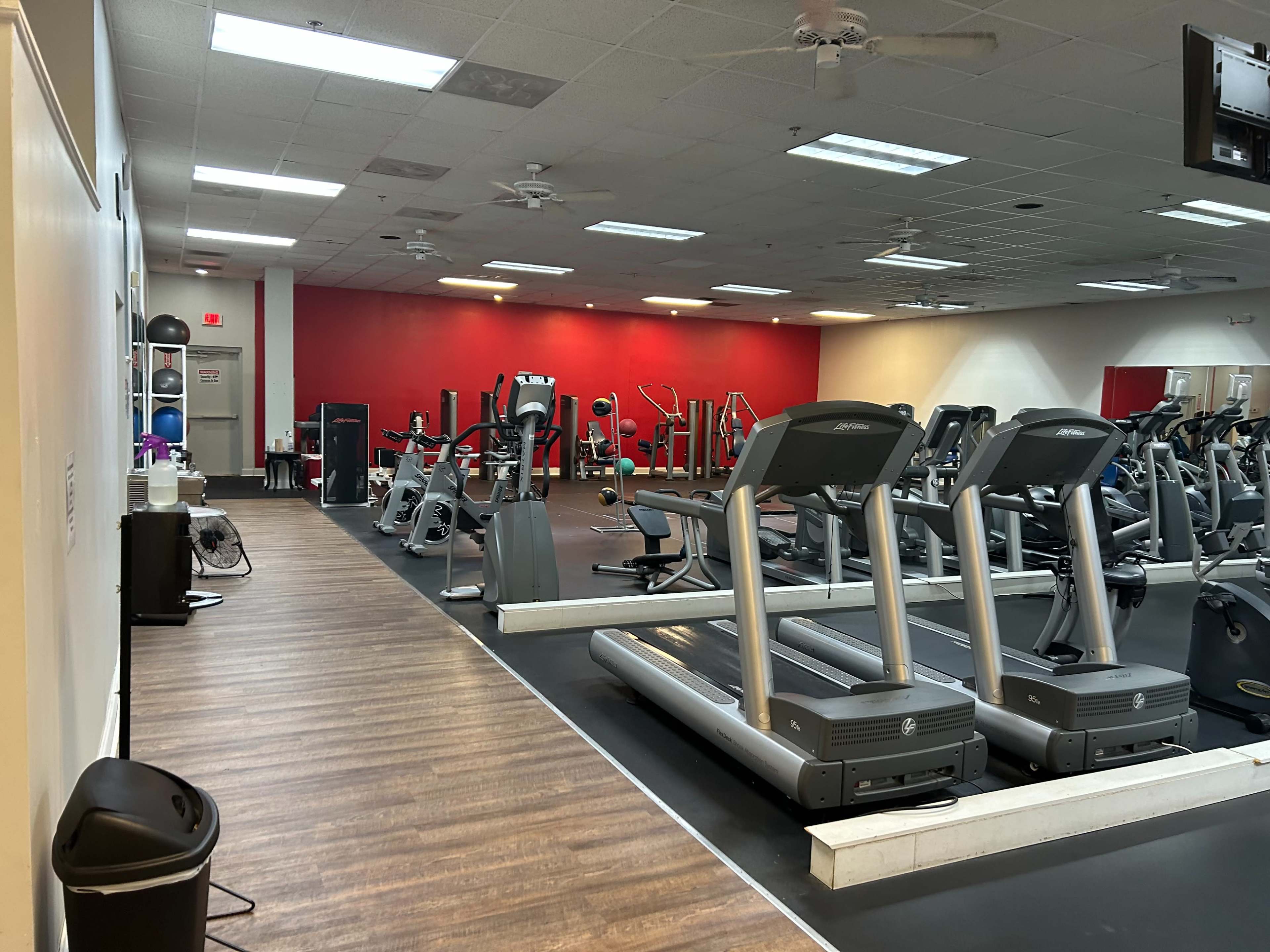 A gym interior with various exercise machines, including treadmills and weights, arranged in an open space with a red accent wall.