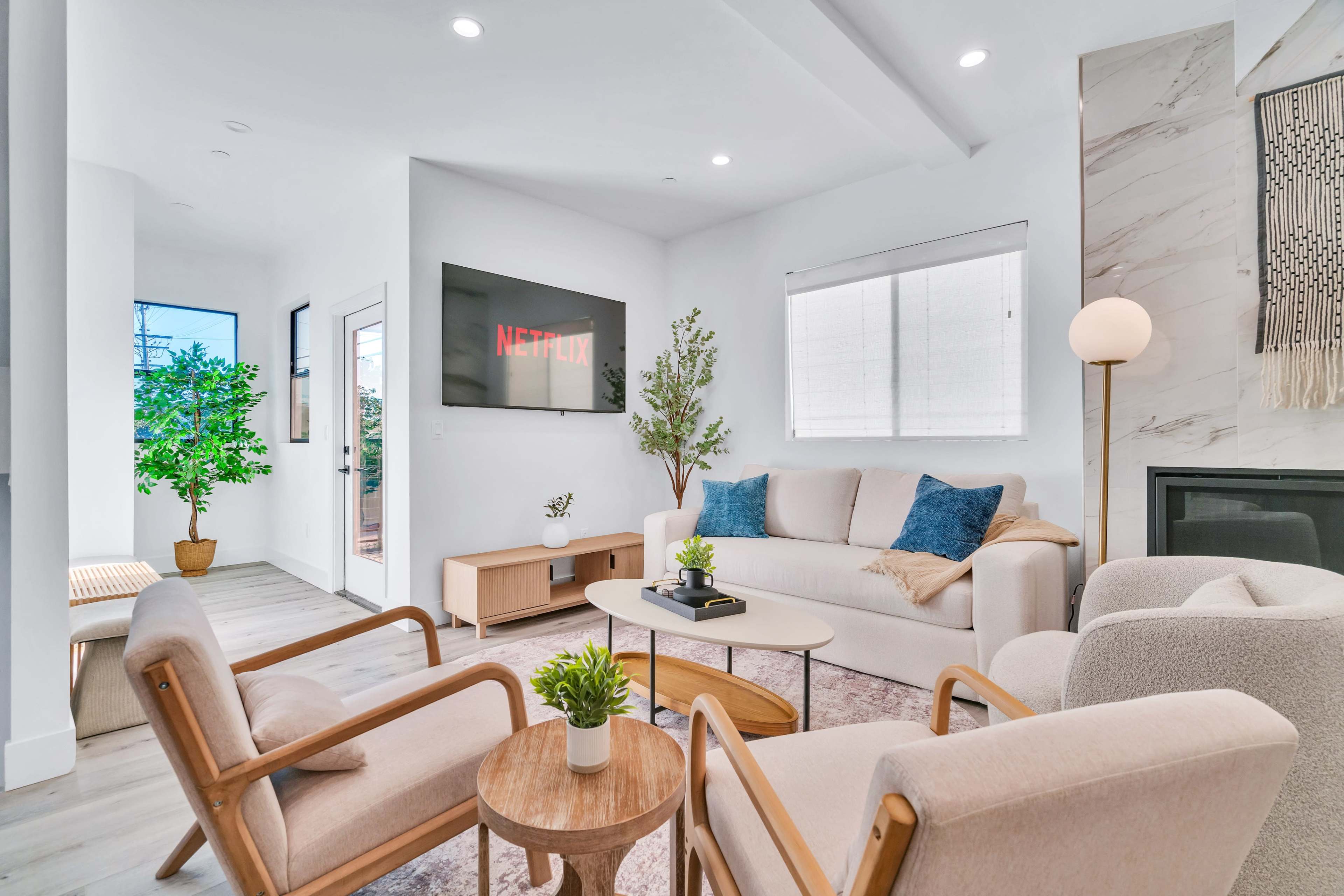 The image shows a modern living room with a light-colored sofa, two armchairs, a round coffee table, and a wall-mounted television displaying the Netflix logo.