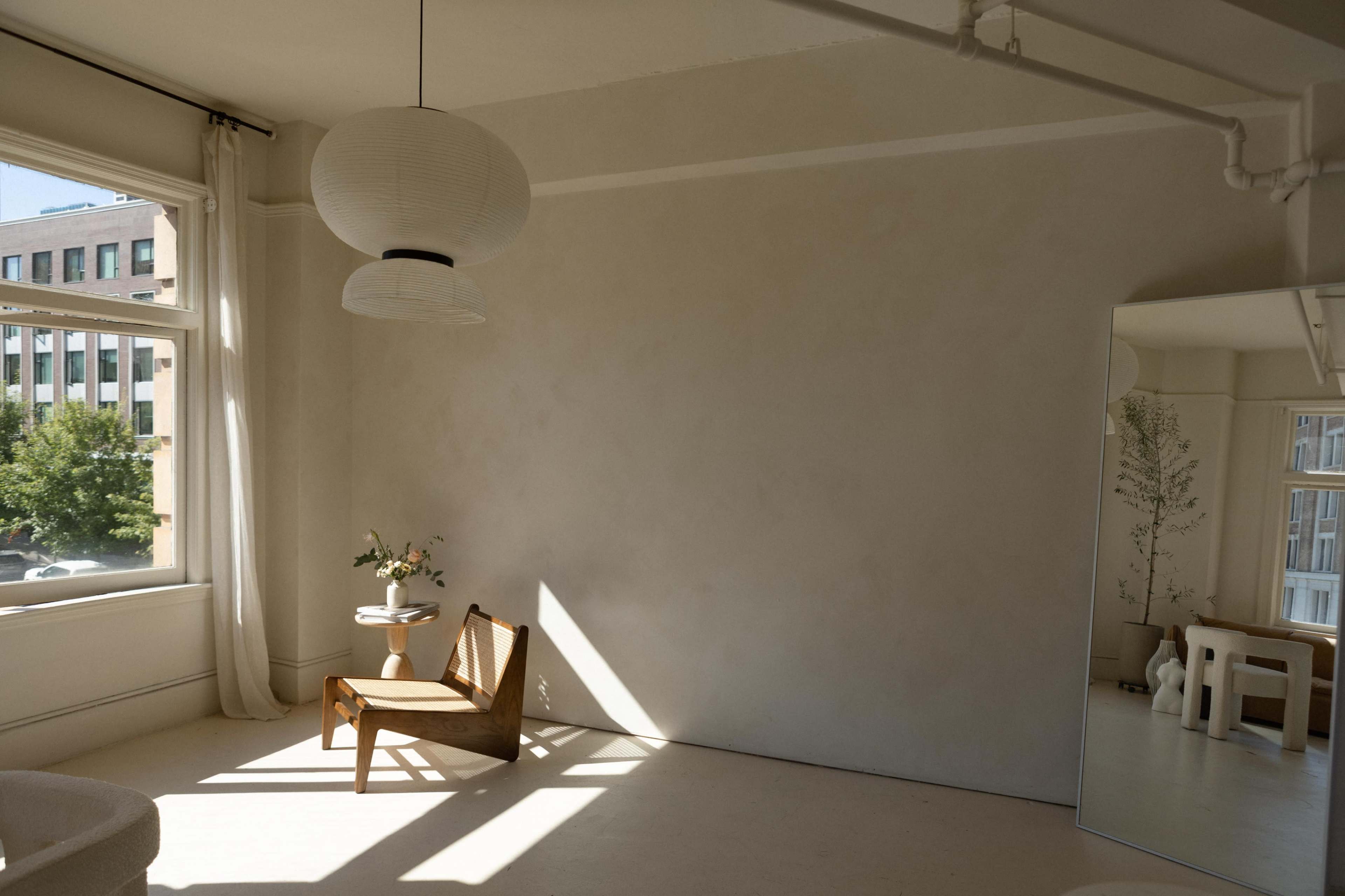 A sunlit room features a simple wooden chair, a small side table with a vase of flowers, and a large mirror reflecting the space.