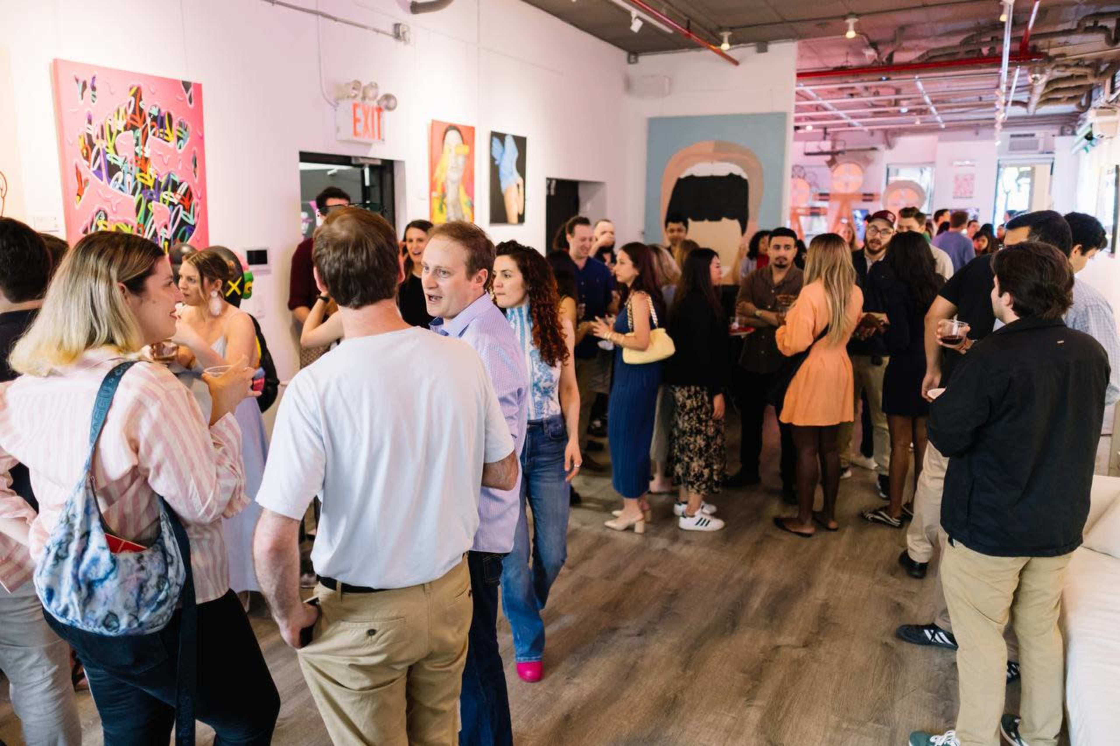A large group of people mingles in a lively art gallery space with colorful artworks displayed on the walls.
