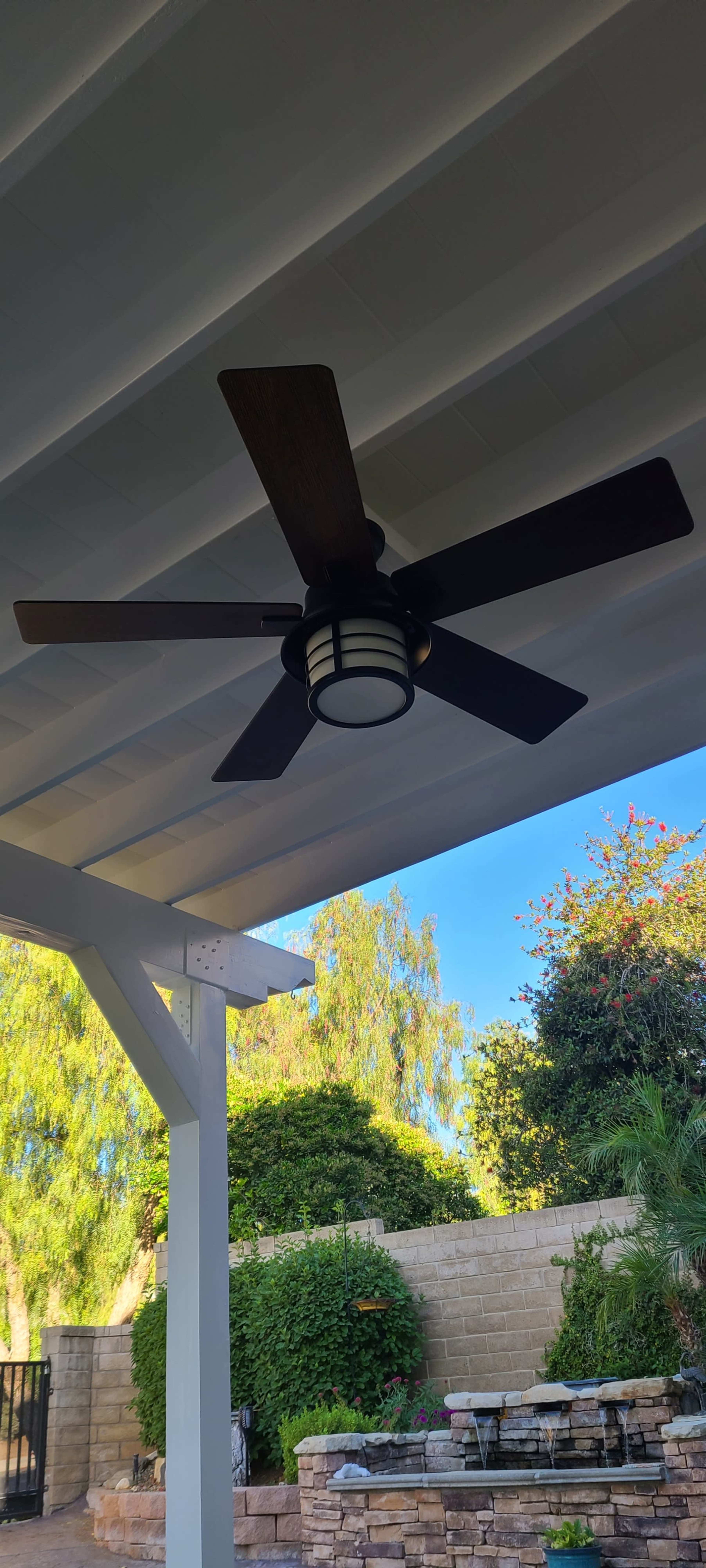 A ceiling fan with five wooden blades and a light fixture is mounted under a covered patio surrounded by greenery.