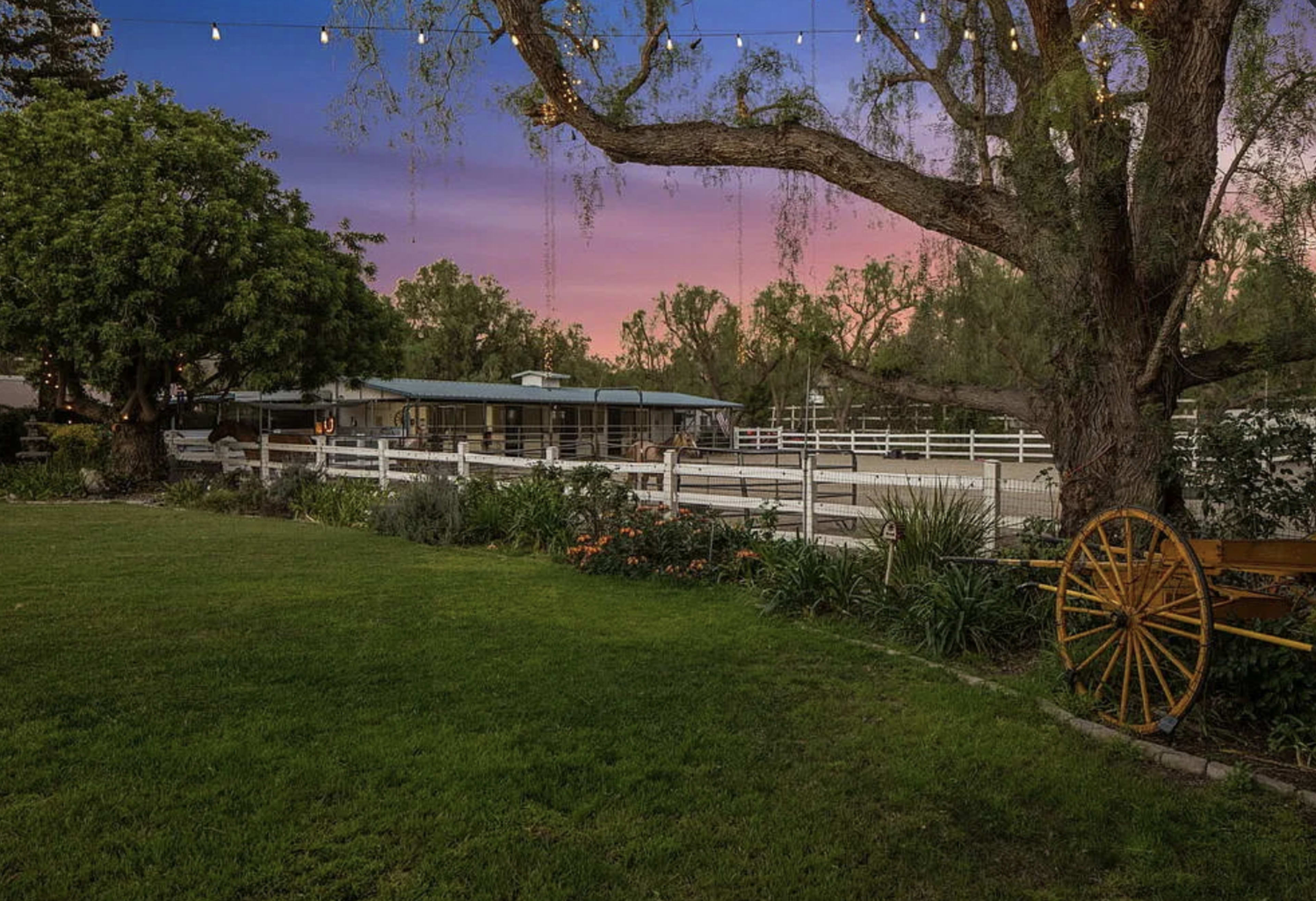 The image shows a grassy area with a large tree and a wooden cart wheel, overlooking a ranch-style building and a fenced riding area under a colorful sky.