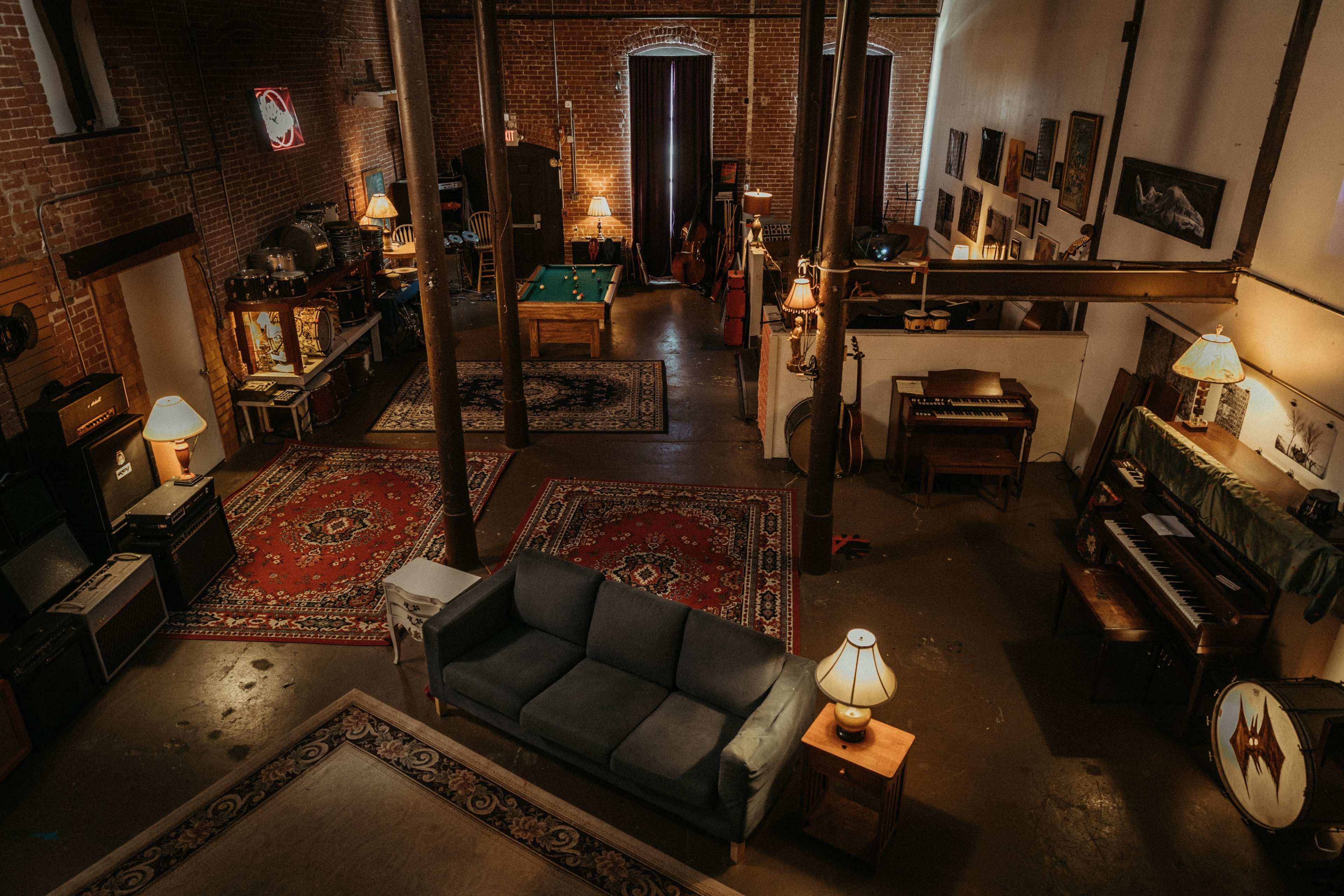 An industrial-style interior features a spacious room with brick walls, a pool table, musical instruments, and various furniture arranged around rugs and lamps.