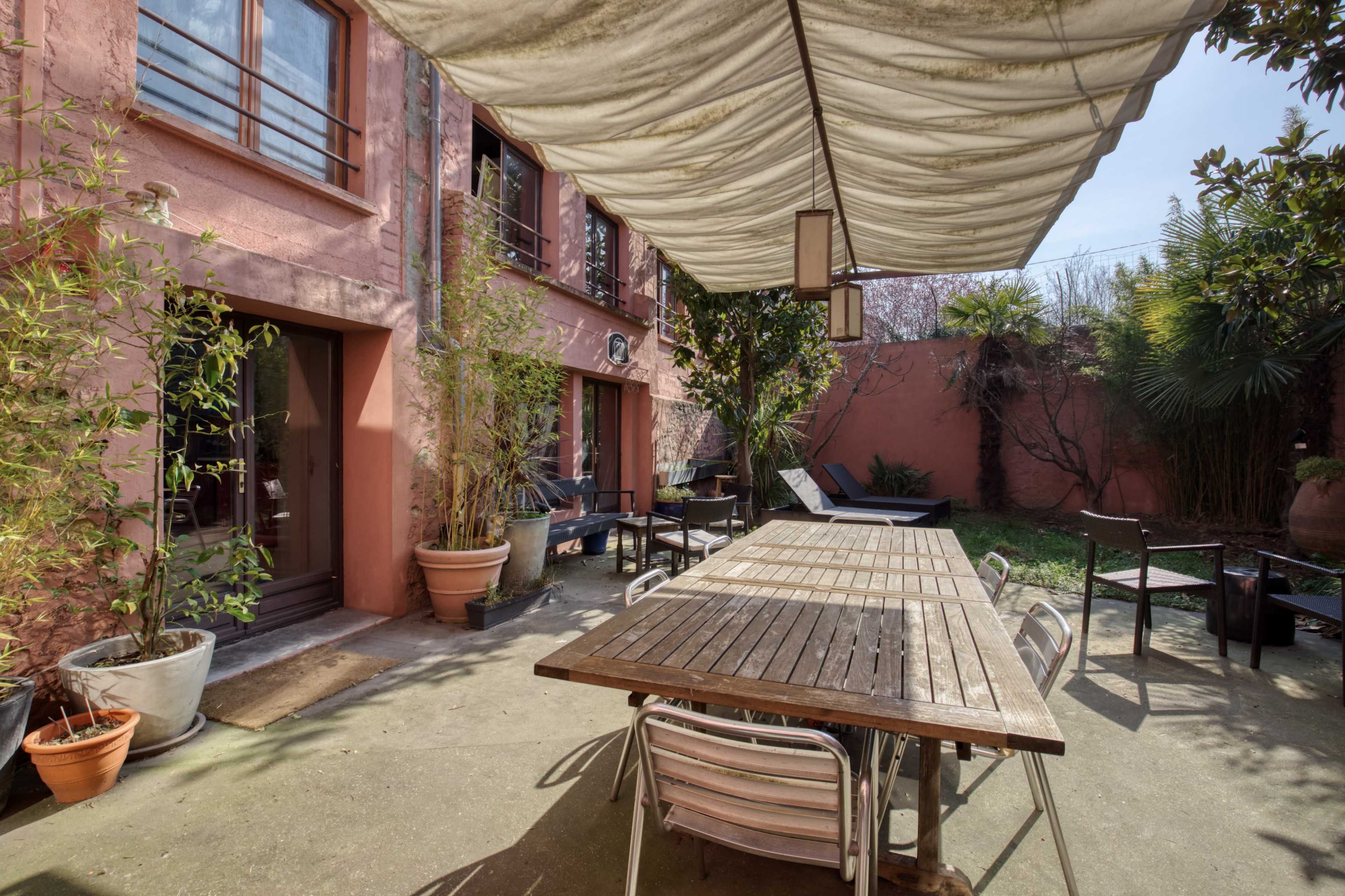 A courtyard with a wooden dining table and chairs beneath a canopy, surrounded by potted plants and buildings with pink walls.