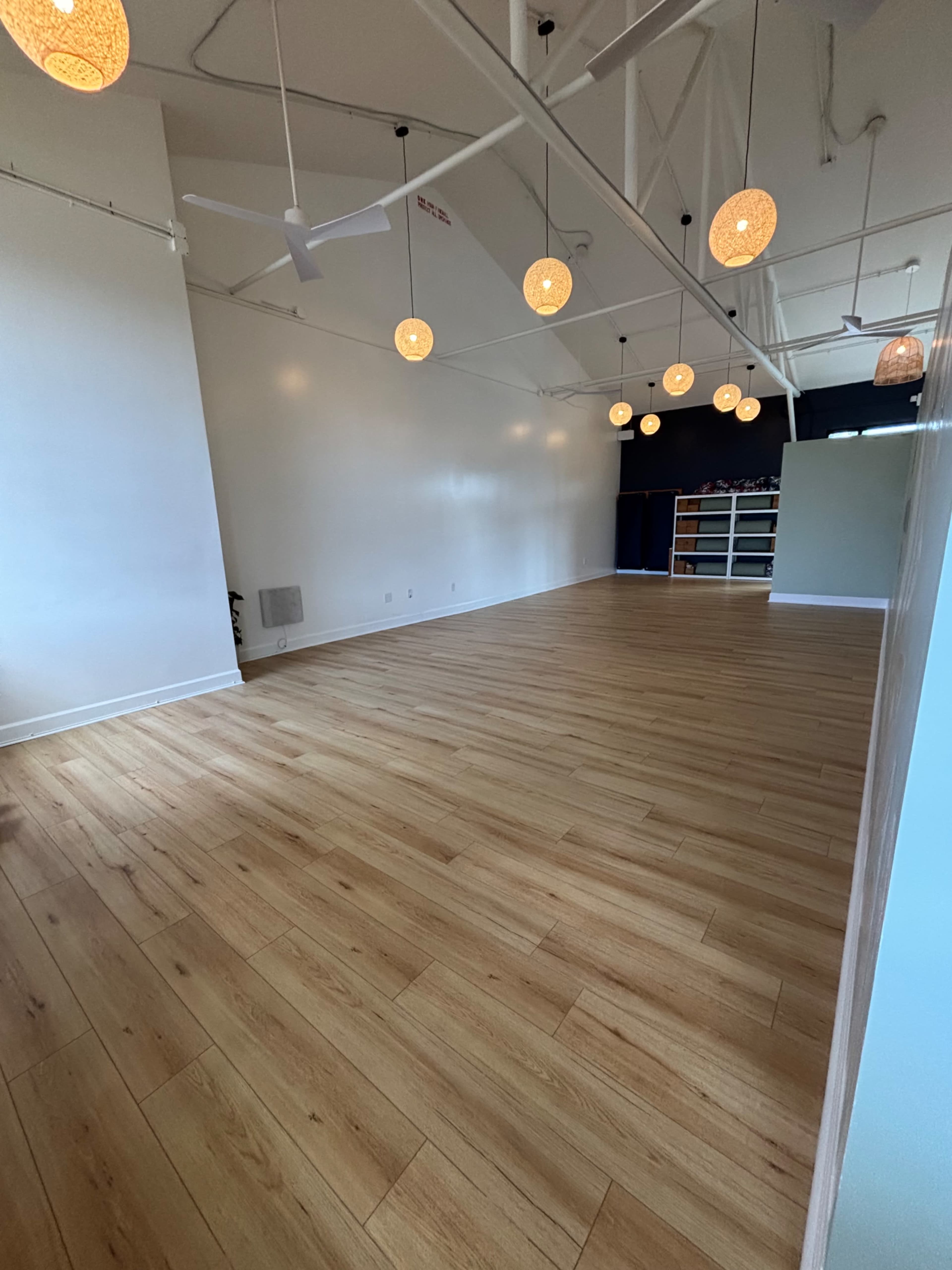 The image depicts a spacious interior with wooden flooring, white walls, and pendant lights hanging from the ceiling.