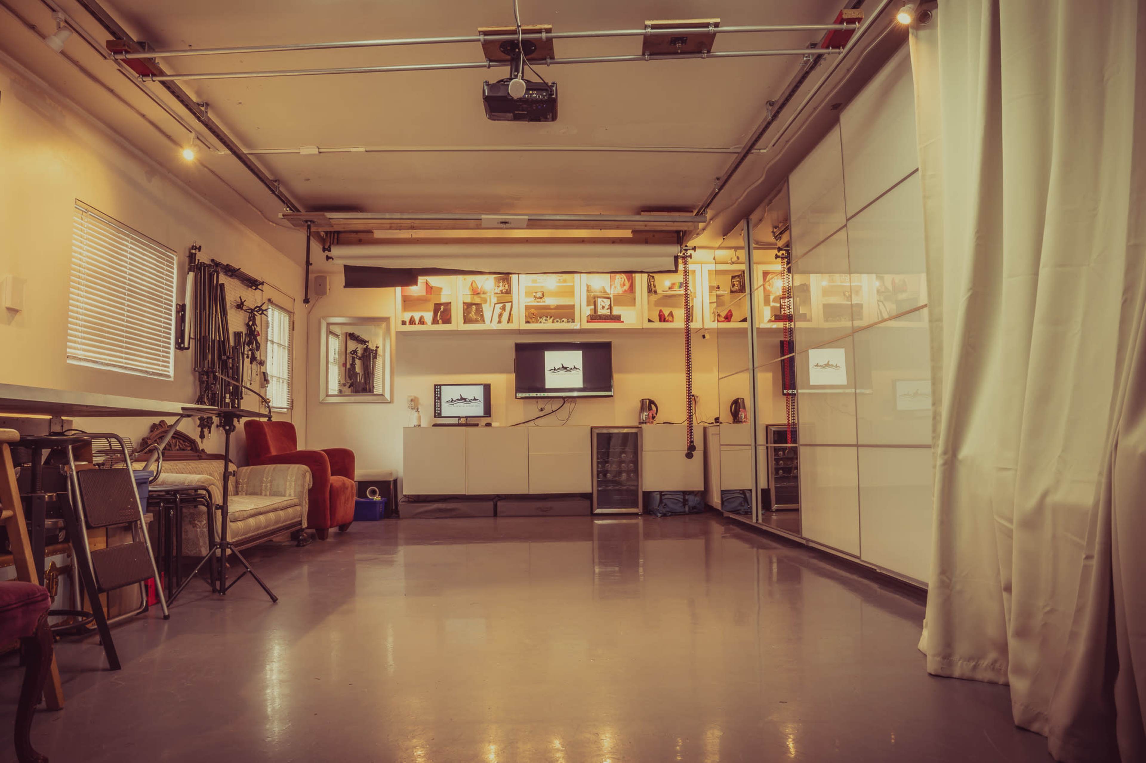 A spacious indoor garage area with a polished floor, fitted with shelves displaying various items, a projector on the ceiling, and two televisions mounted on a wall.