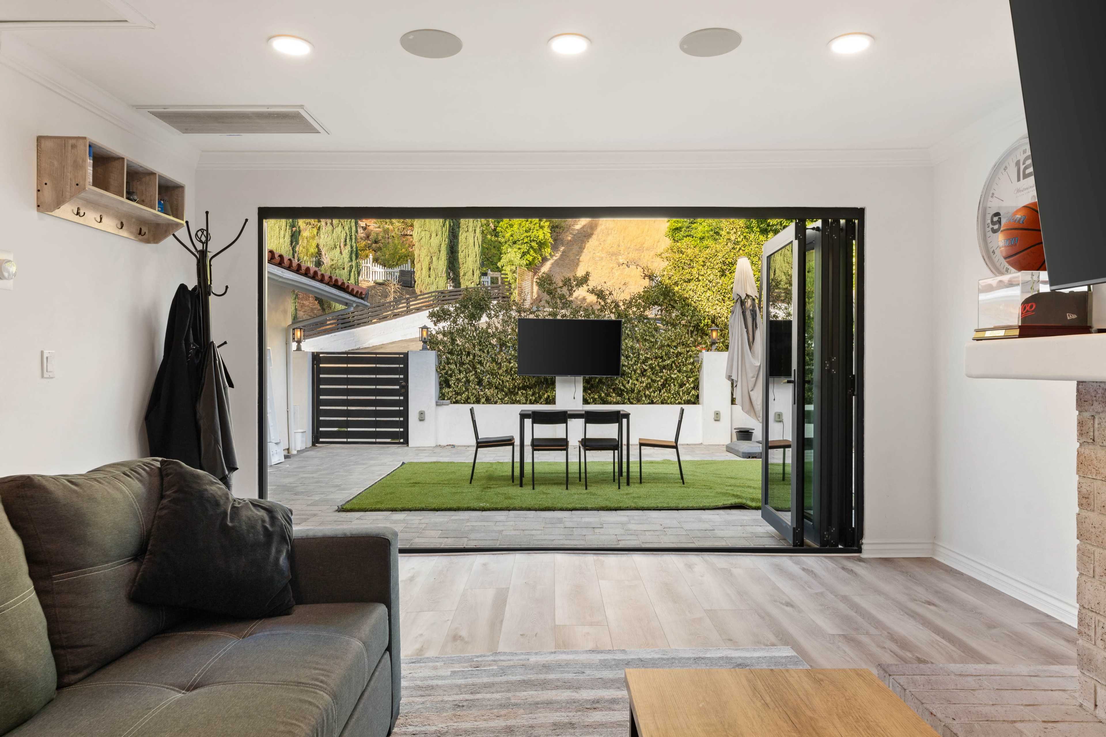 The image shows a modern living room that opens up to a patio area with outdoor furniture and a television set against a backdrop of greenery.
