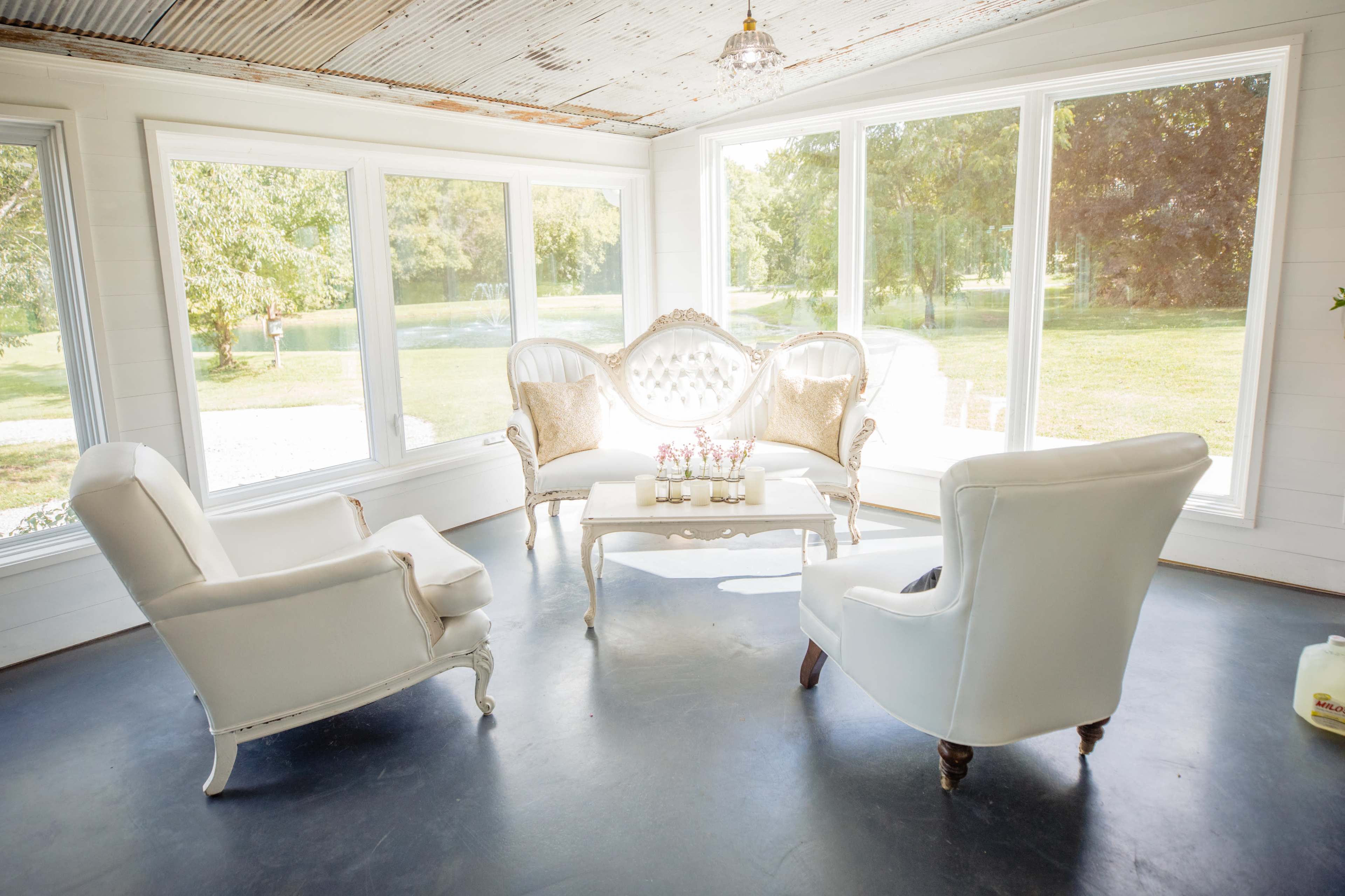 The image shows a bright, airy room with large windows, featuring a white upholstered set of chairs and a decorative sofa arranged around a central coffee table.