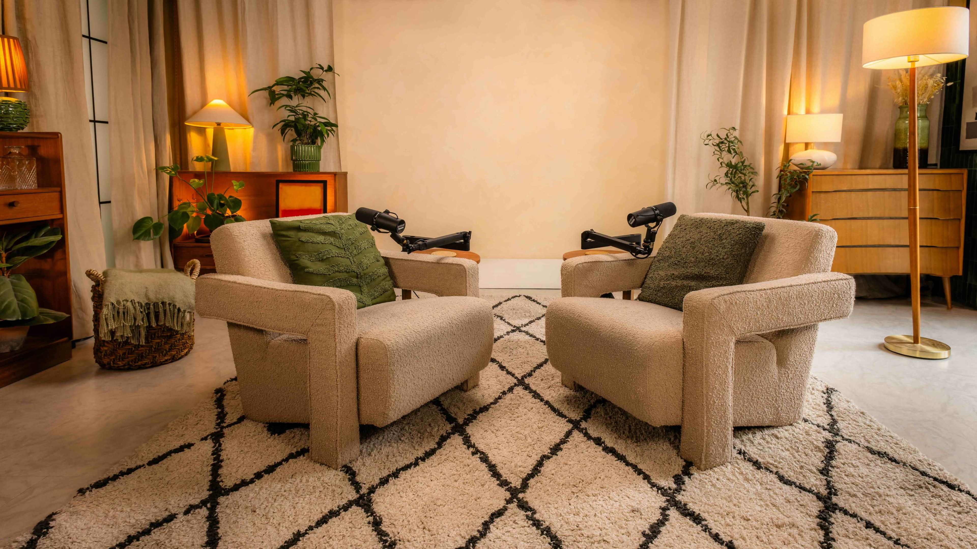 The image shows a cozy interior scene with two upholstered armchairs facing each other, surrounded by plants and warm lighting.