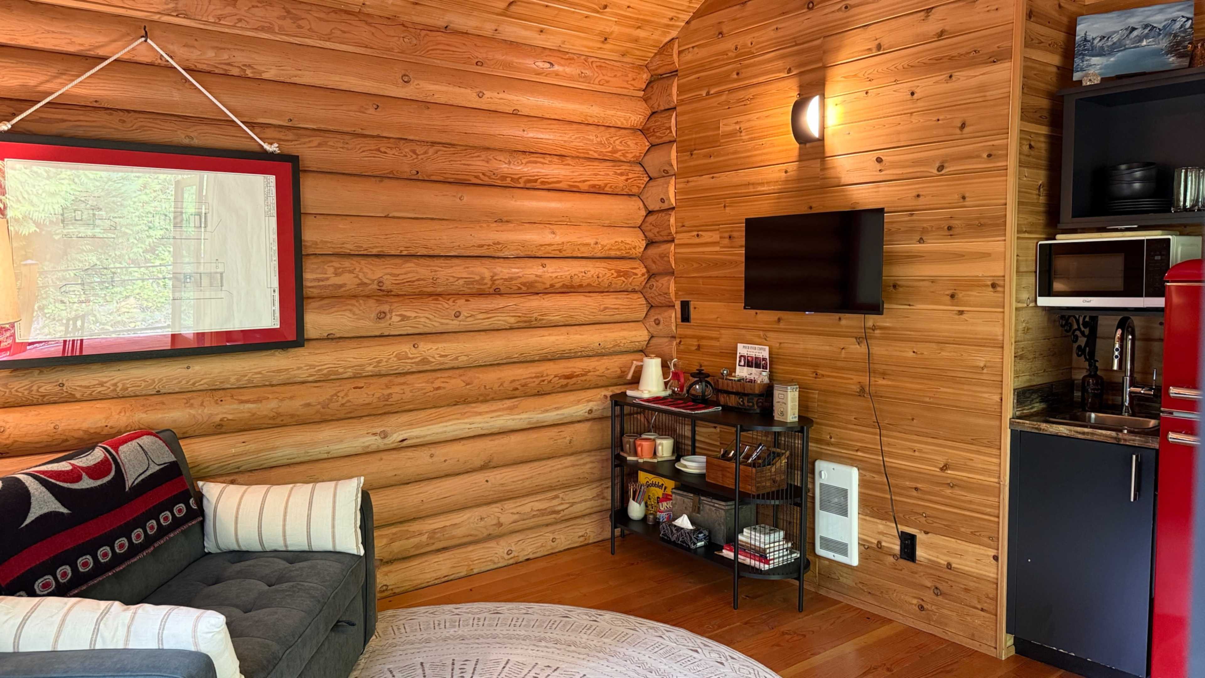 The interior of a log cabin features a cozy seating area with a gray sofa, a wall-mounted TV, and a small kitchen space with a red refrigerator and various kitchen appliances.
