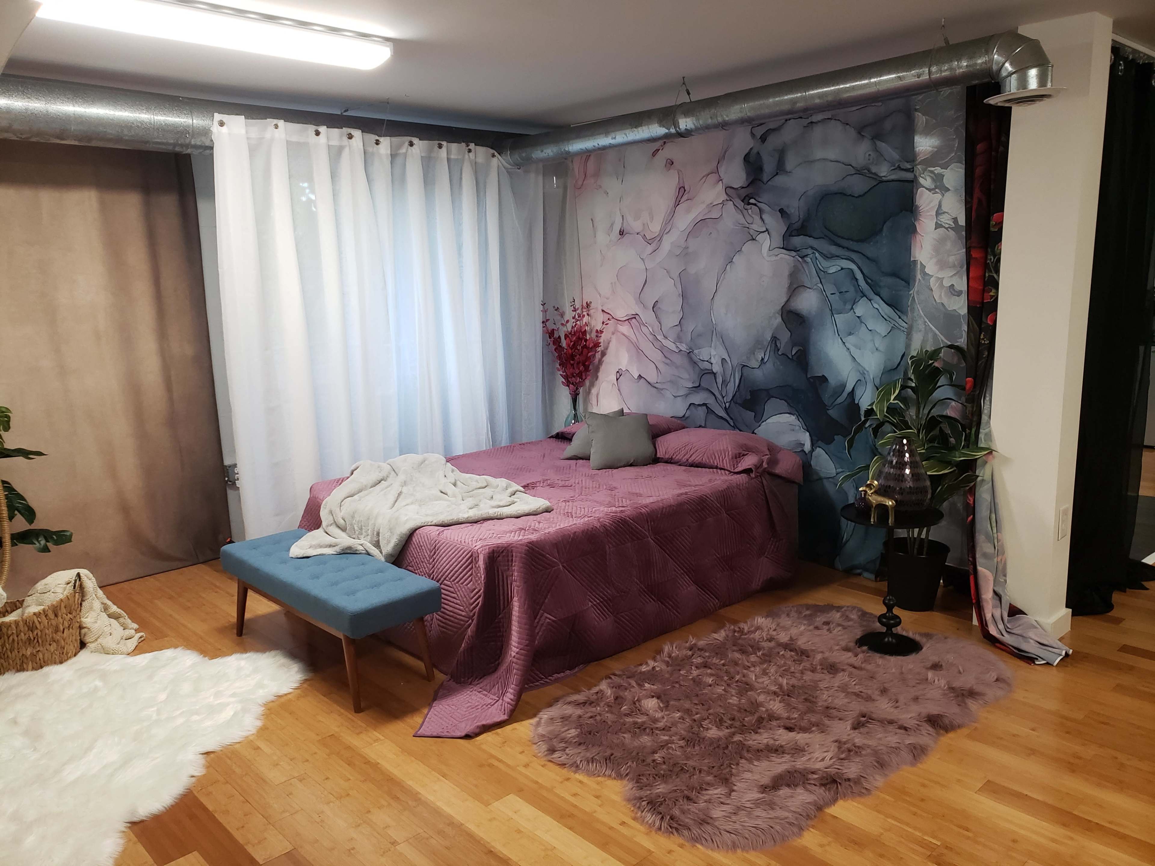 The image shows a bedroom with a bed covered in a purple quilt, a blue bench at the foot, and a colorful abstract mural on the wall, complementing a hardwood floor.