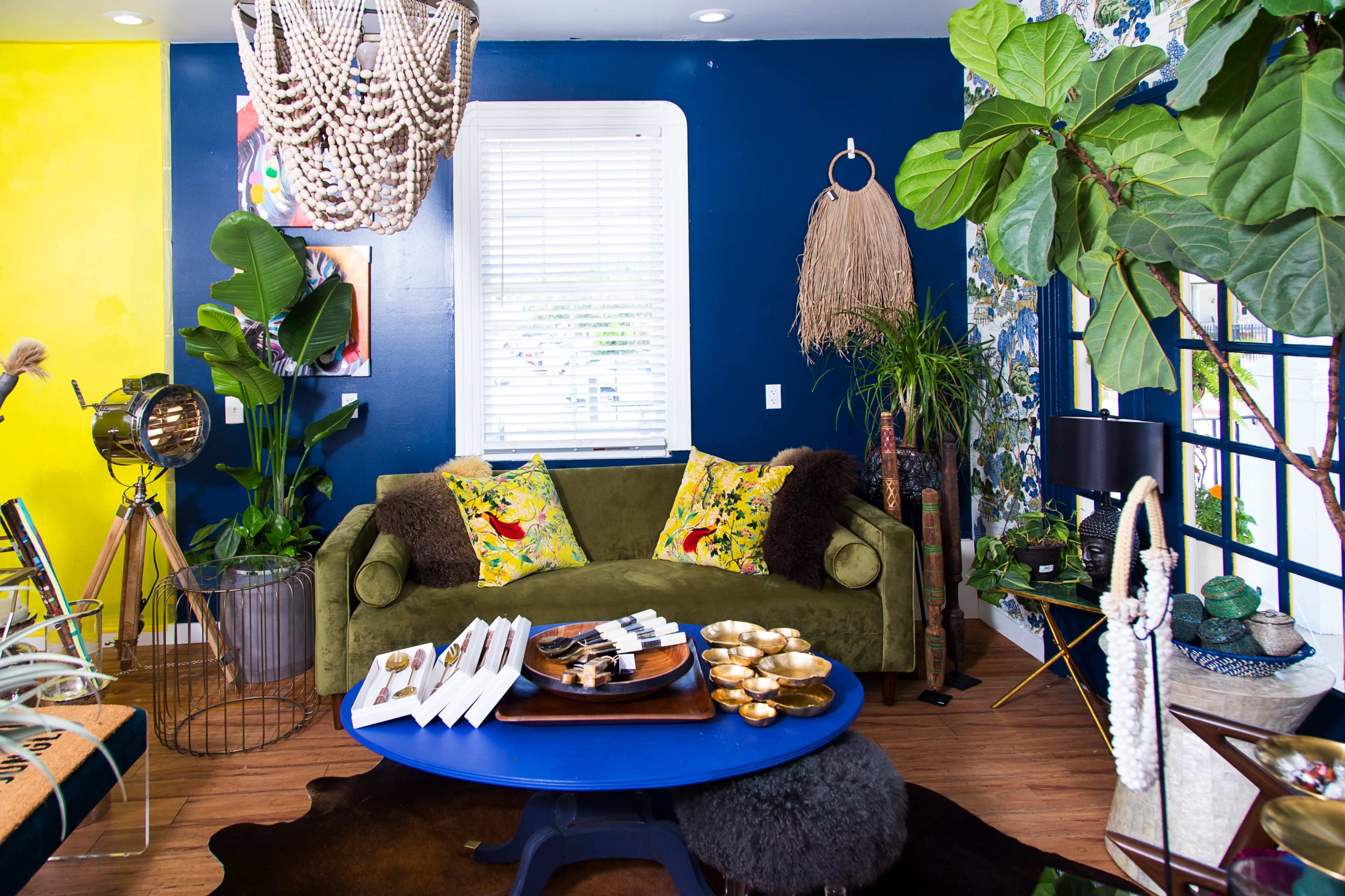 The image shows a vibrant living room with a green sofa, decorative pillows, a circular blue coffee table, and various plants and decorative items against a backdrop of blue and yellow walls.