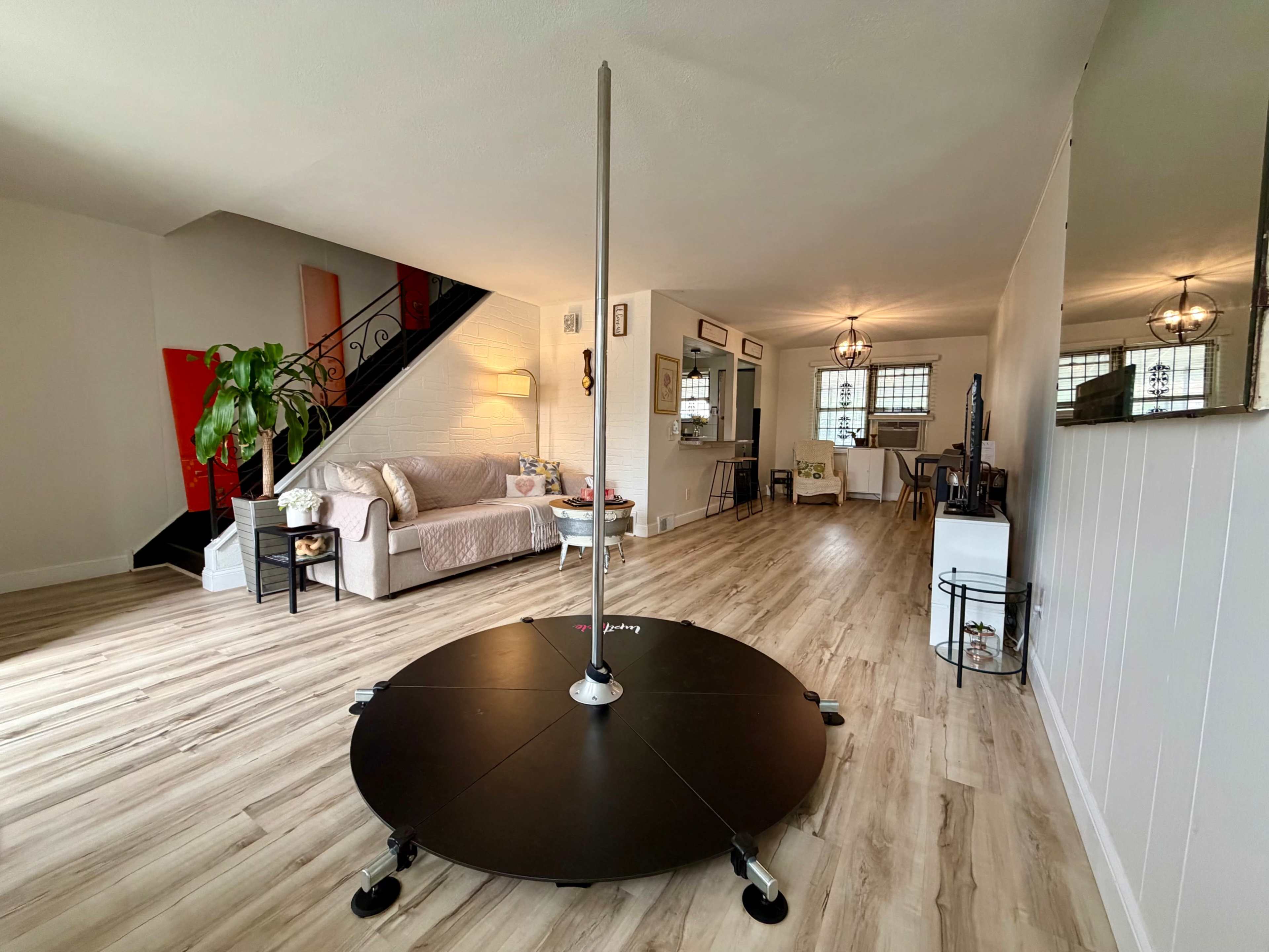 The image shows a spacious living area with a pole dancing setup in the center, featuring wooden flooring and a staircase in the background leading to a second level.