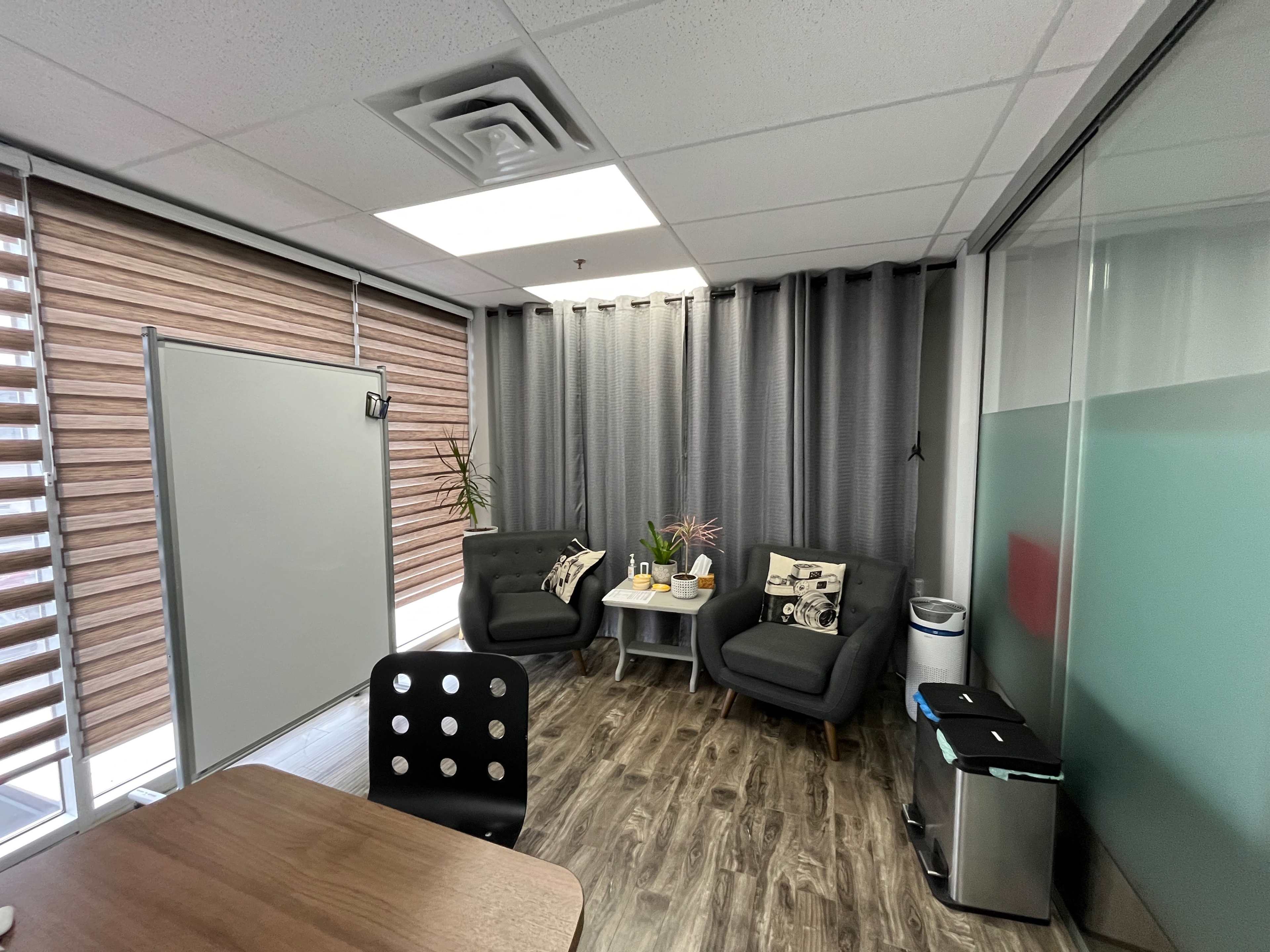 The image shows a modern office space featuring two dark green chairs, a small table, and a glass wall, with blinds partially covering the windows.