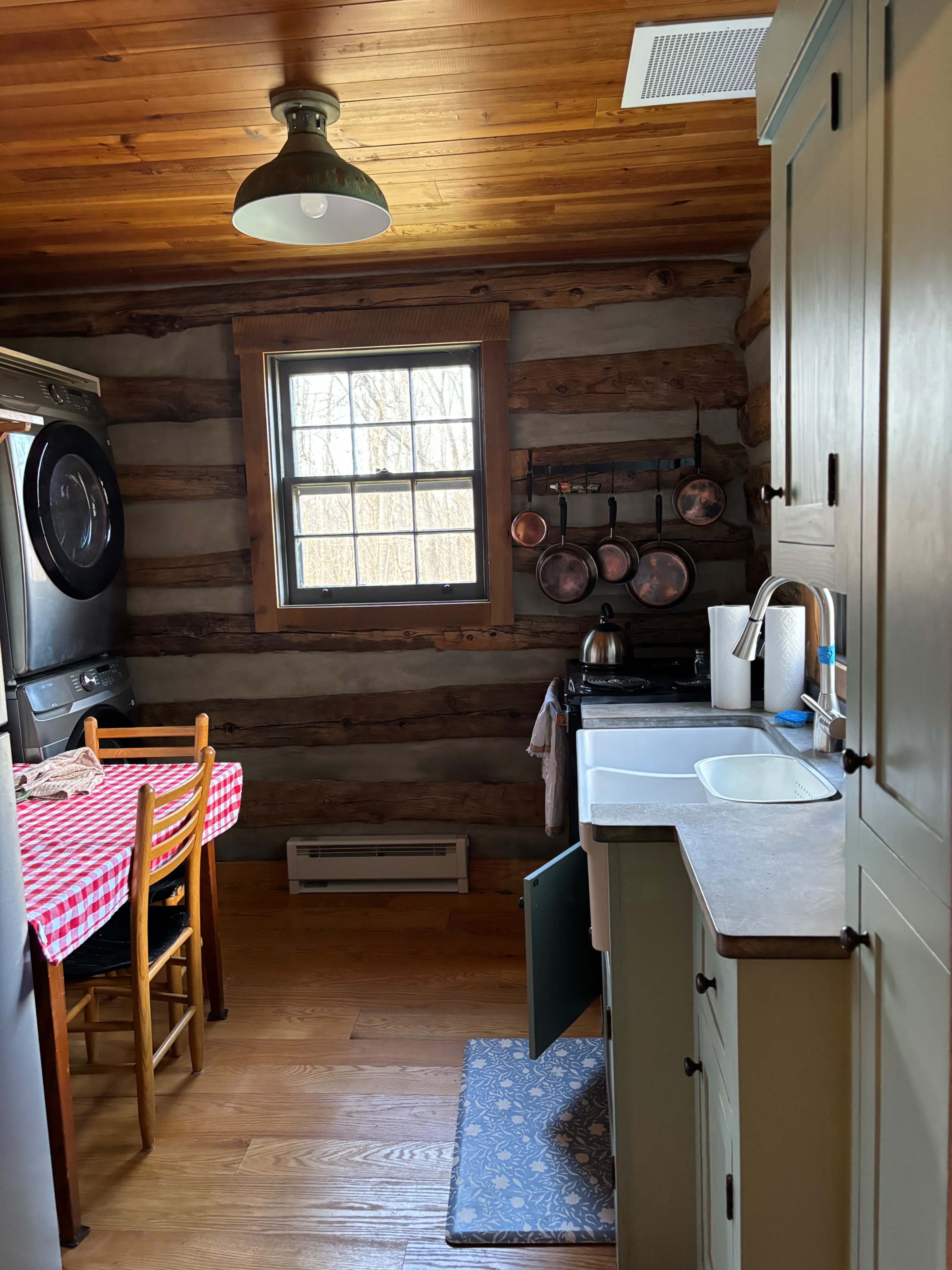 The image shows a rustic kitchen with wooden log walls, a table covered in a red and white checkered cloth, and copper cookware hanging above a stove near a window.
