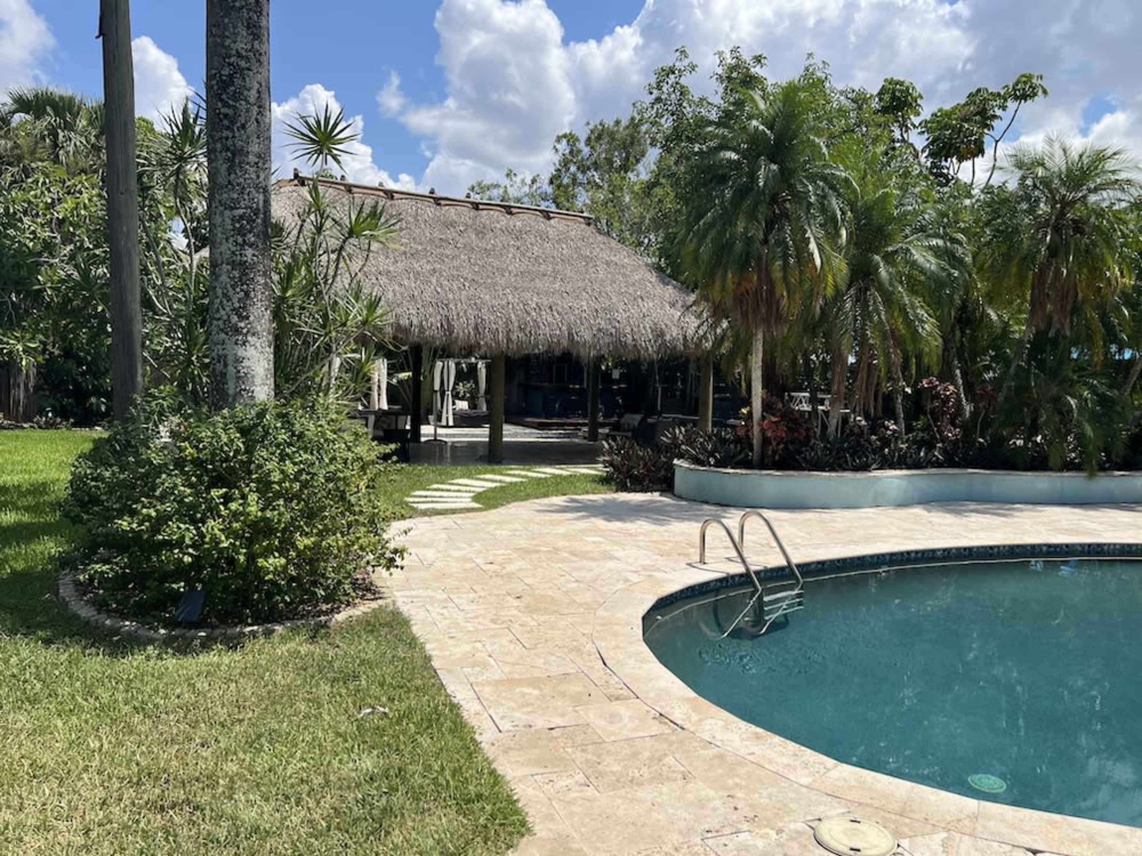 A thatched-roof structure is situated beside a swimming pool, surrounded by lush greenery and palm trees.