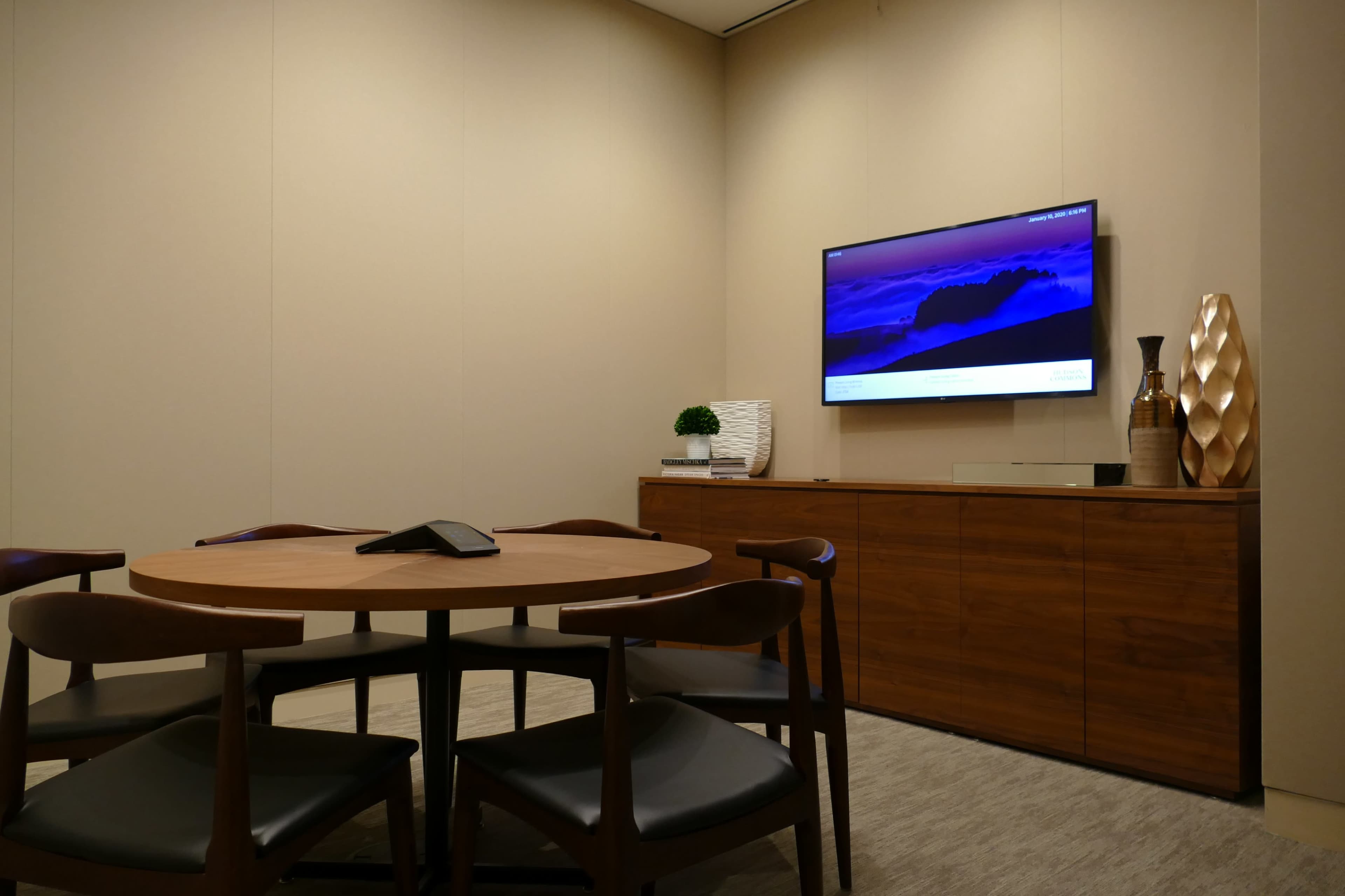 A small conference room features a round table with black chairs, a wooden sideboard, and a wall-mounted television displaying a landscape image.