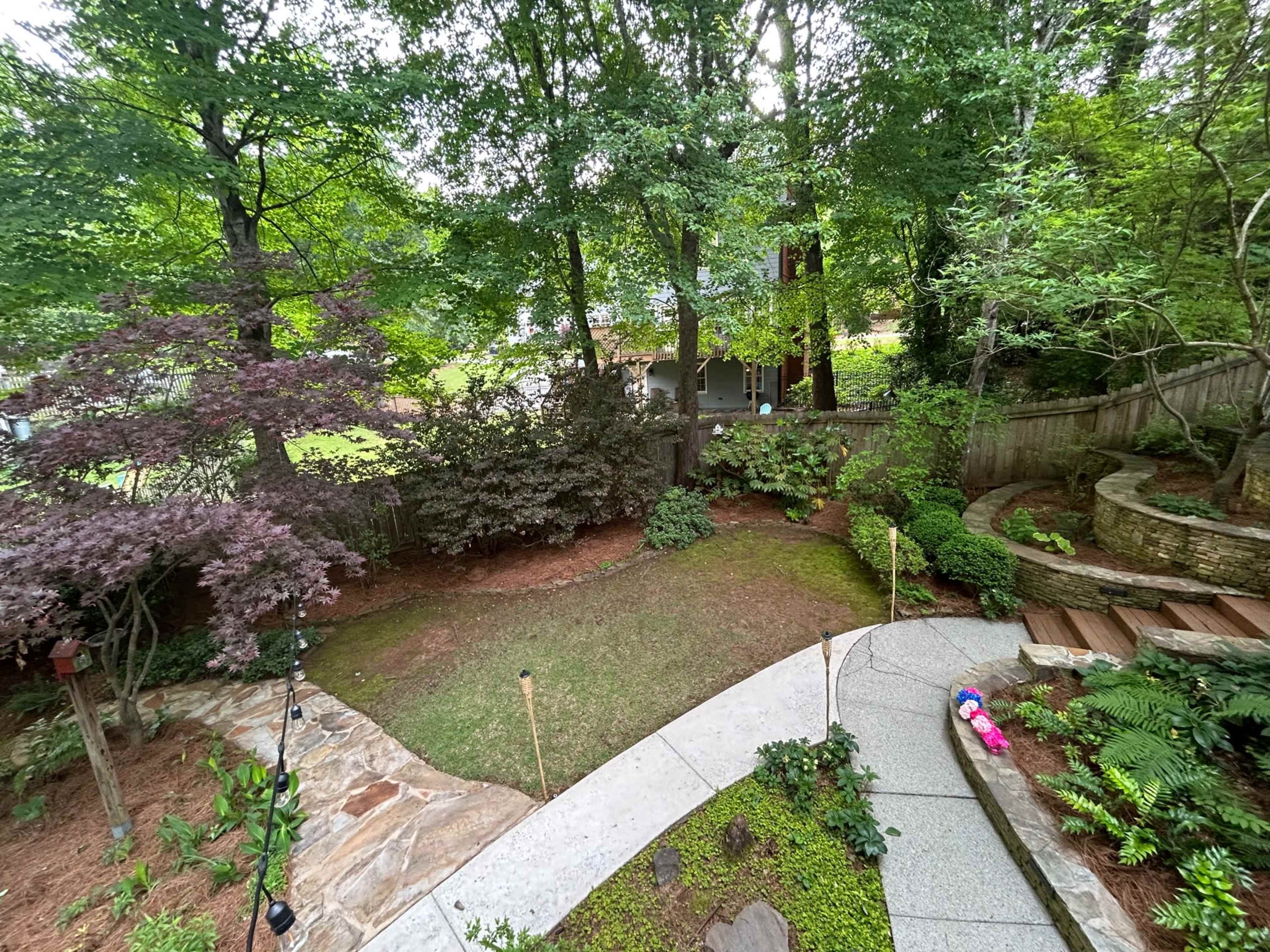 The image shows a landscaped backyard featuring stone pathways, patches of grass, trees, and a wooden deck.