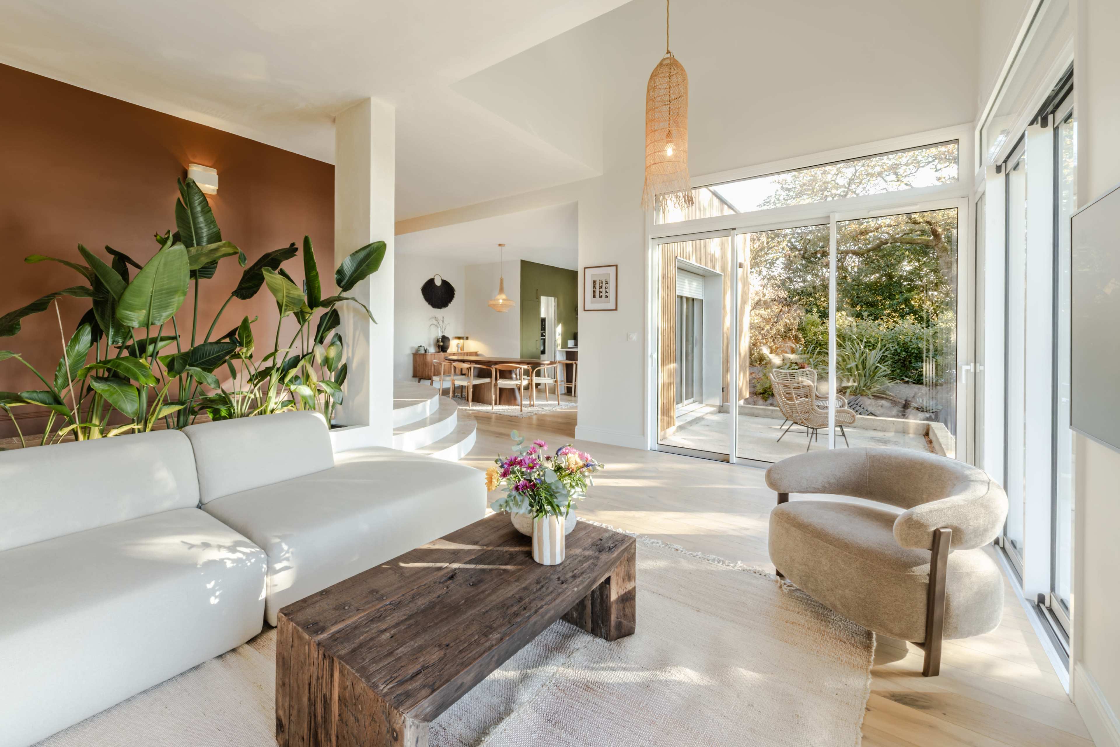 The image shows a modern living room with a light-colored sofa, a wooden coffee table, and large glass doors leading to an outdoor space with plants.