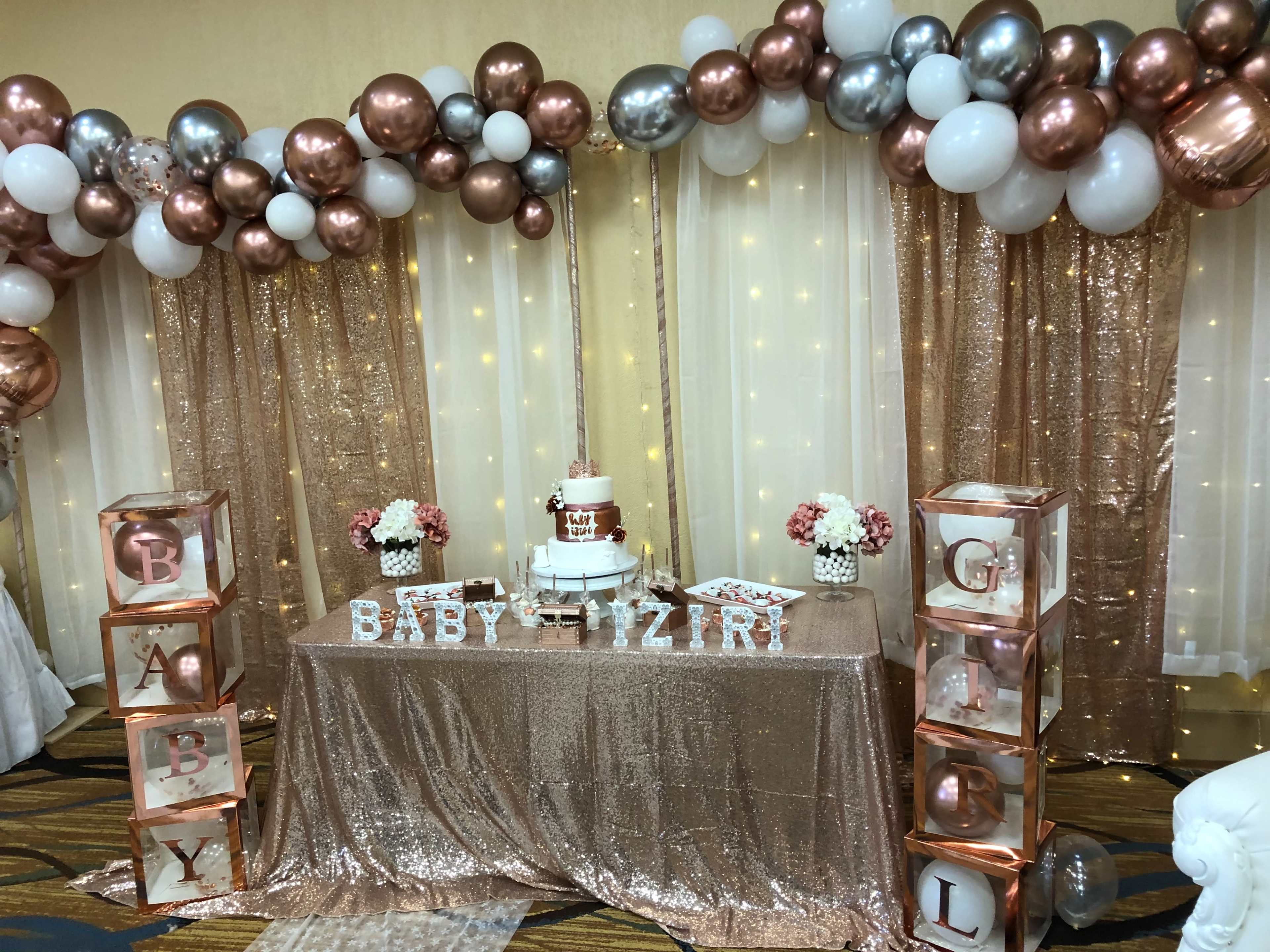 A decorated event space with a table covered in a rose gold sequined cloth, featuring a multi-tiered cake, balloons in various colors above, and letters spelling out "BABY GIRL."