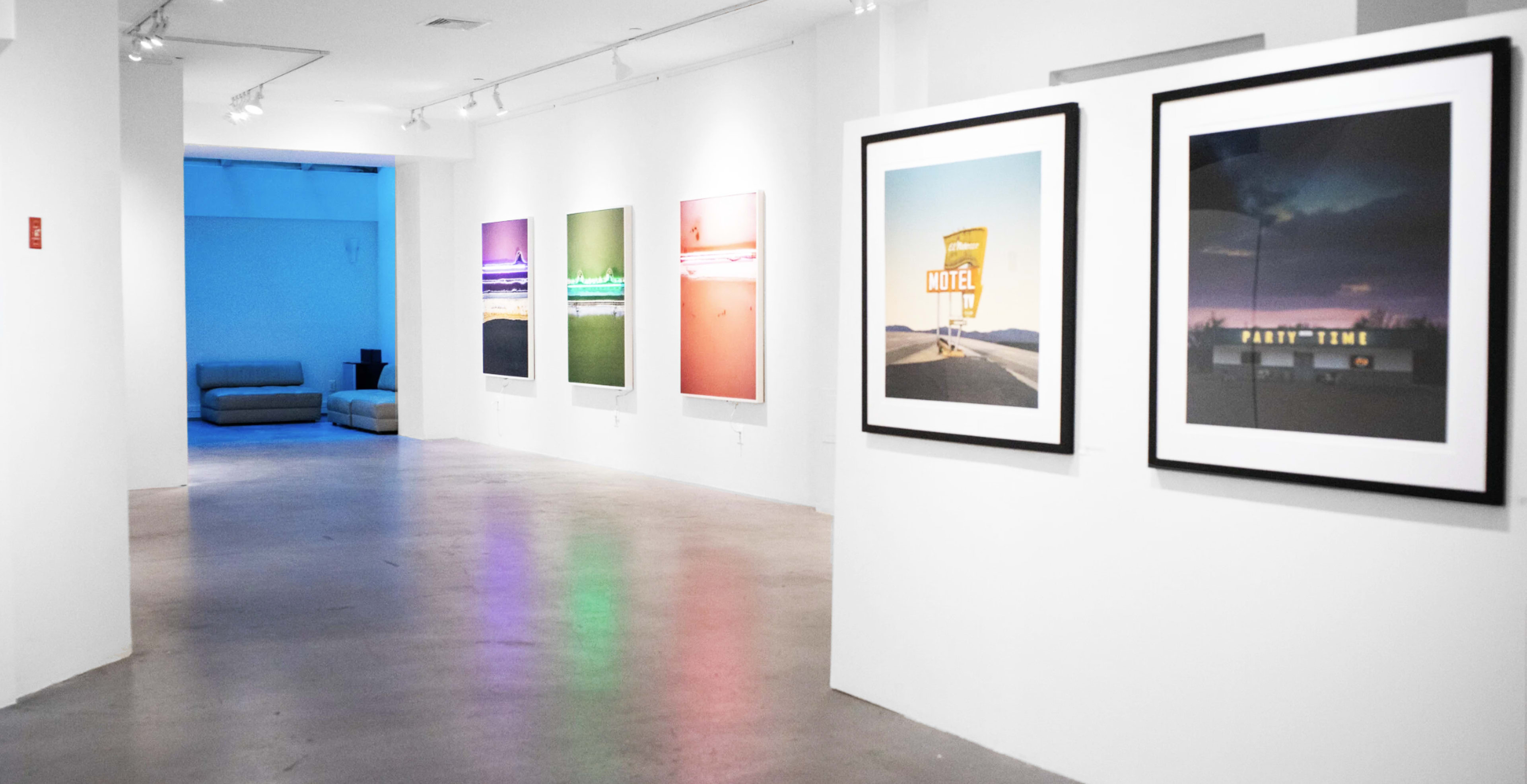 The image shows an art gallery with framed photographs displayed on white walls, leading to a blue-lit area in the background.