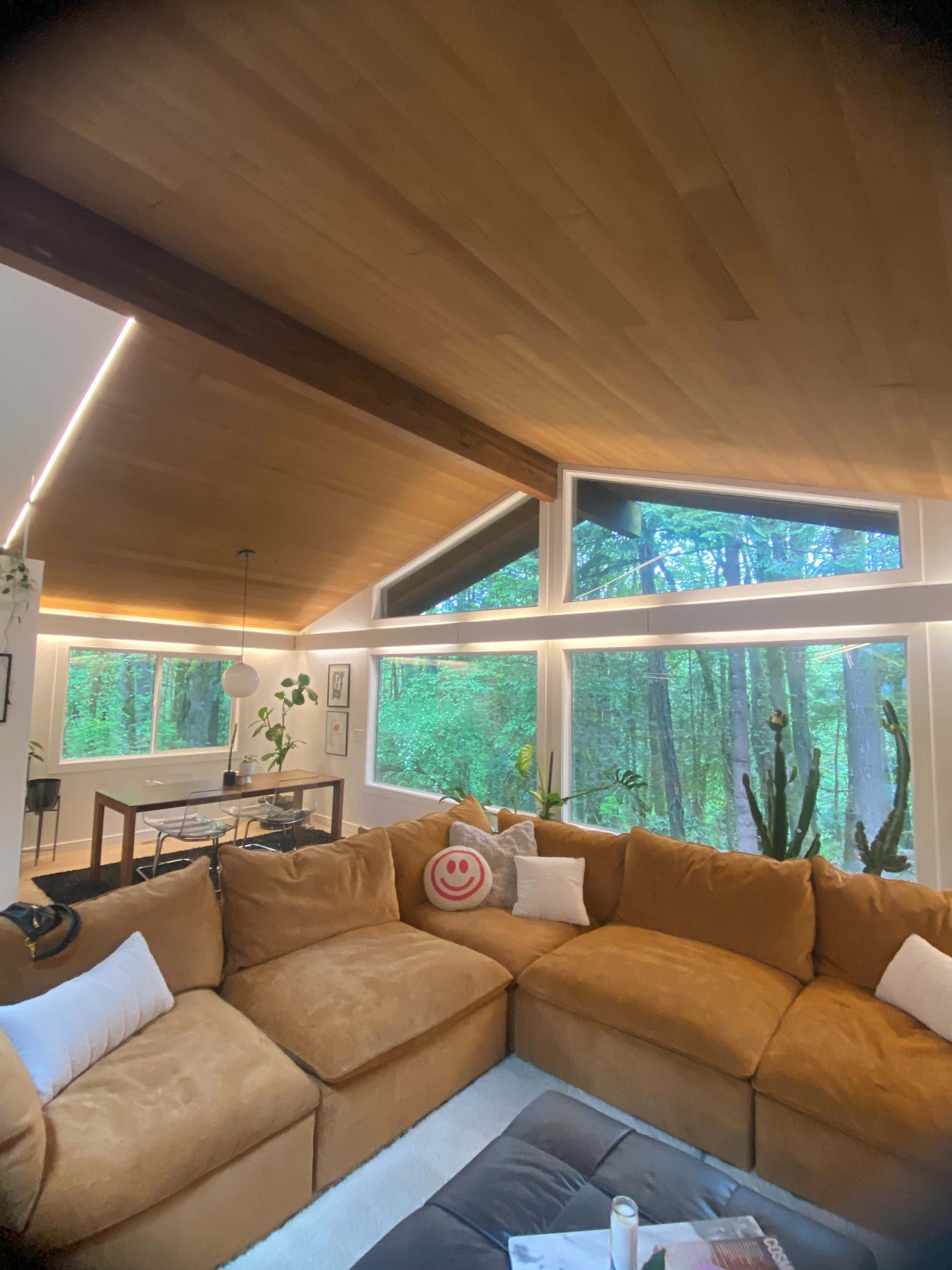 A cozy living room with a large, curved sectional sofa, wooden ceiling, and large windows overlooking a wooded area.