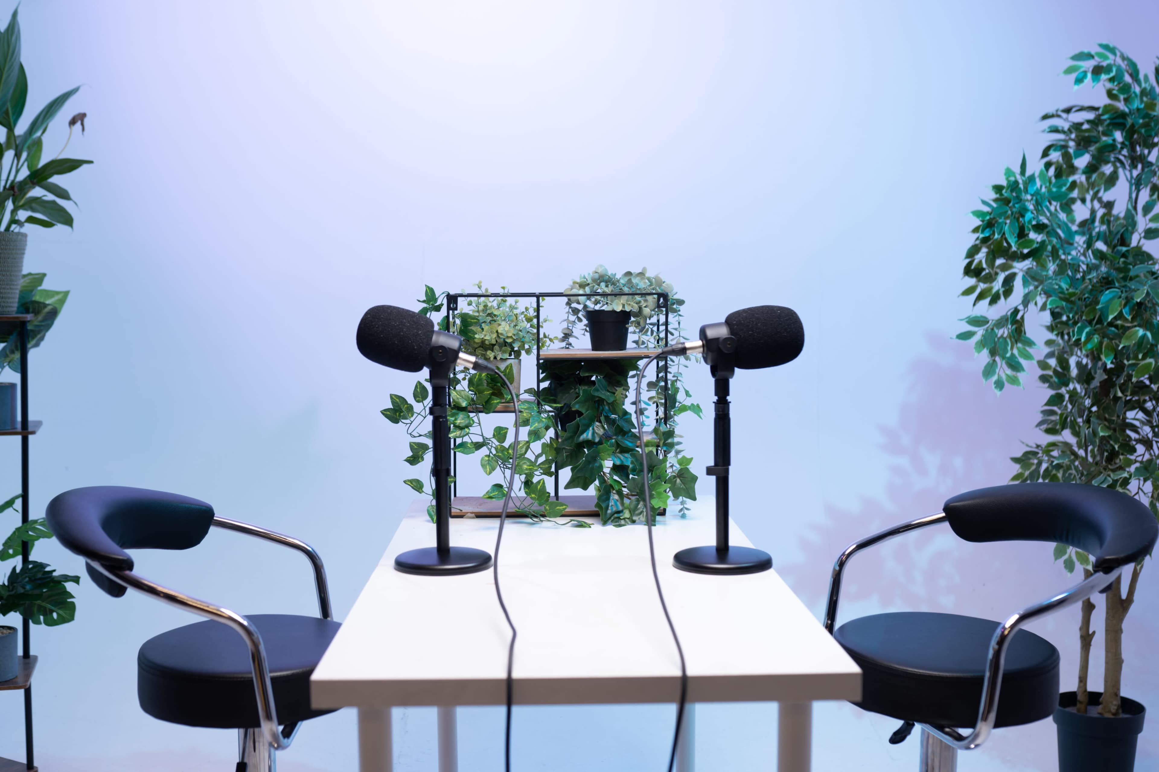 A minimalist podcast studio features a white table with two microphones and chairs, surrounded by greenery against a softly lit background.