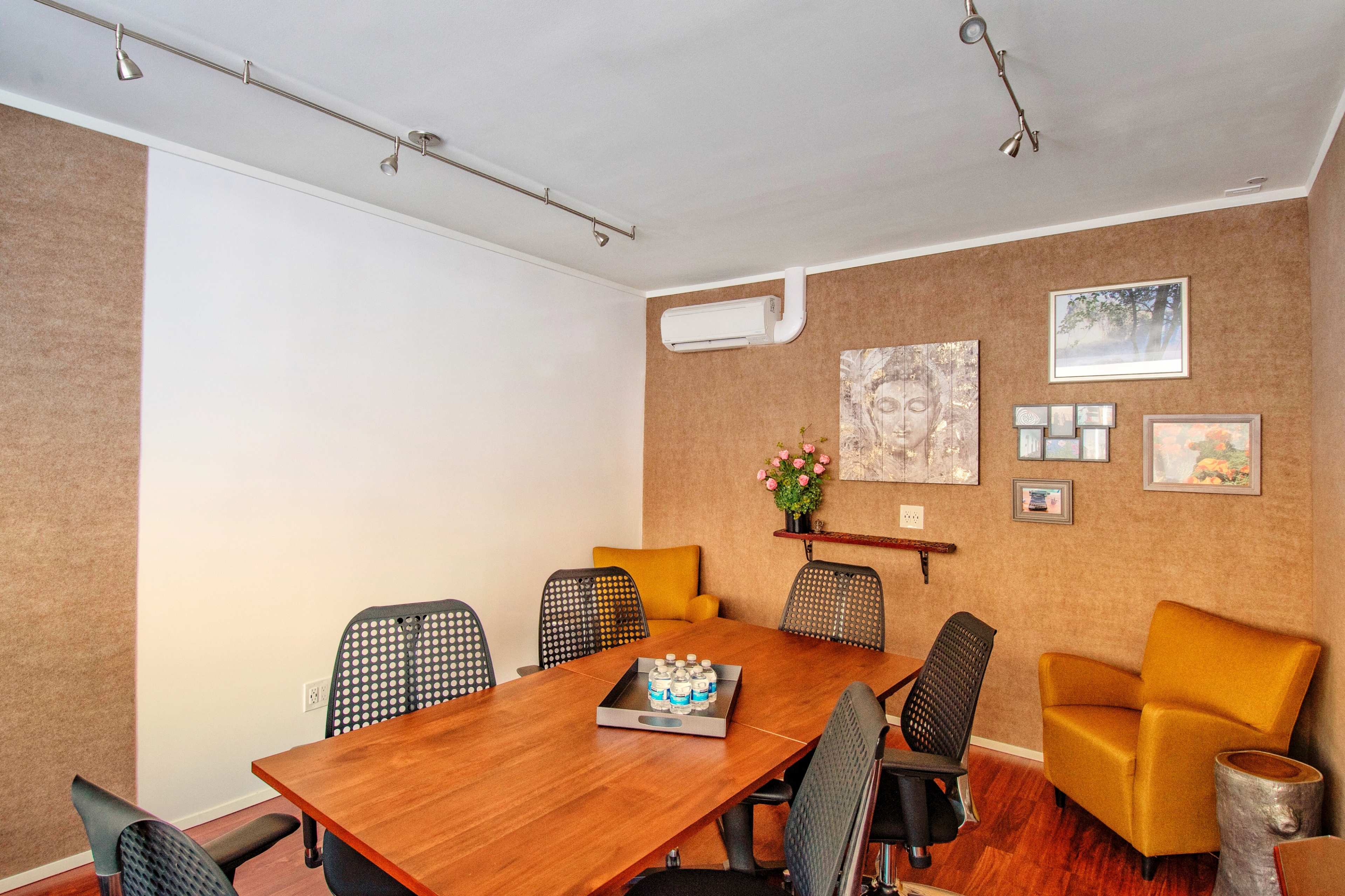 A conference room features a wooden table surrounded by black mesh chairs, with two yellow armchairs and artwork on the walls.