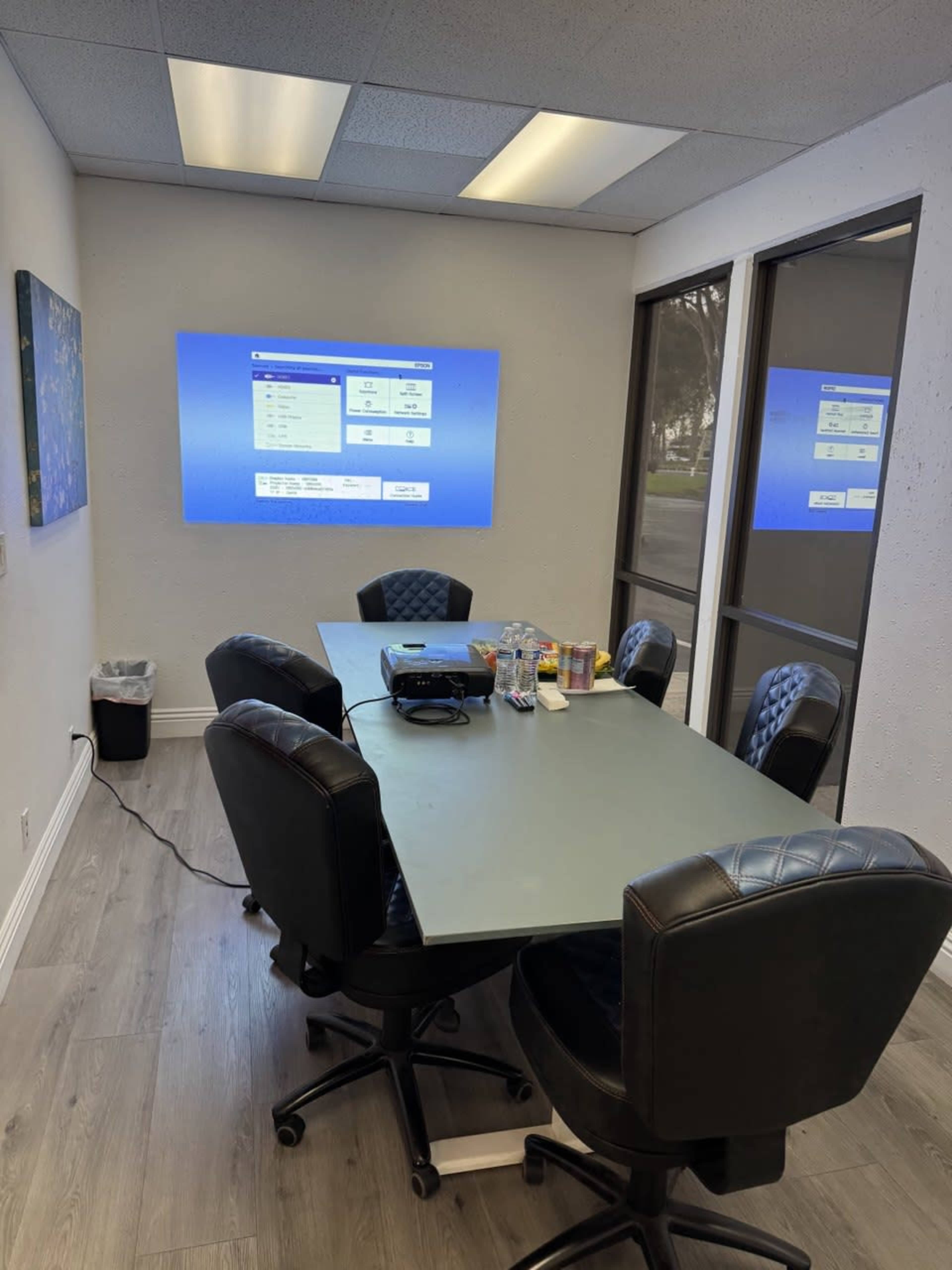 The image shows a conference room with a rectangular table surrounded by six chairs, featuring a projector displaying a digital screen on the wall.