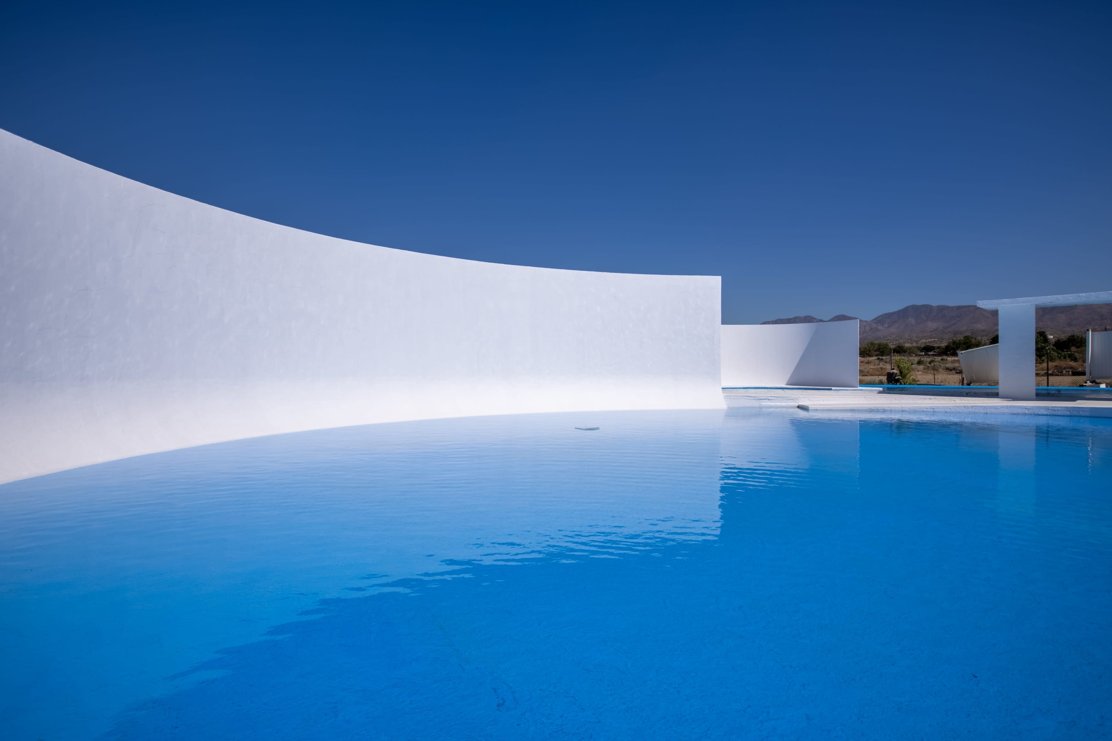 The image features a bright blue swimming pool surrounded by white curved walls under a clear blue sky.