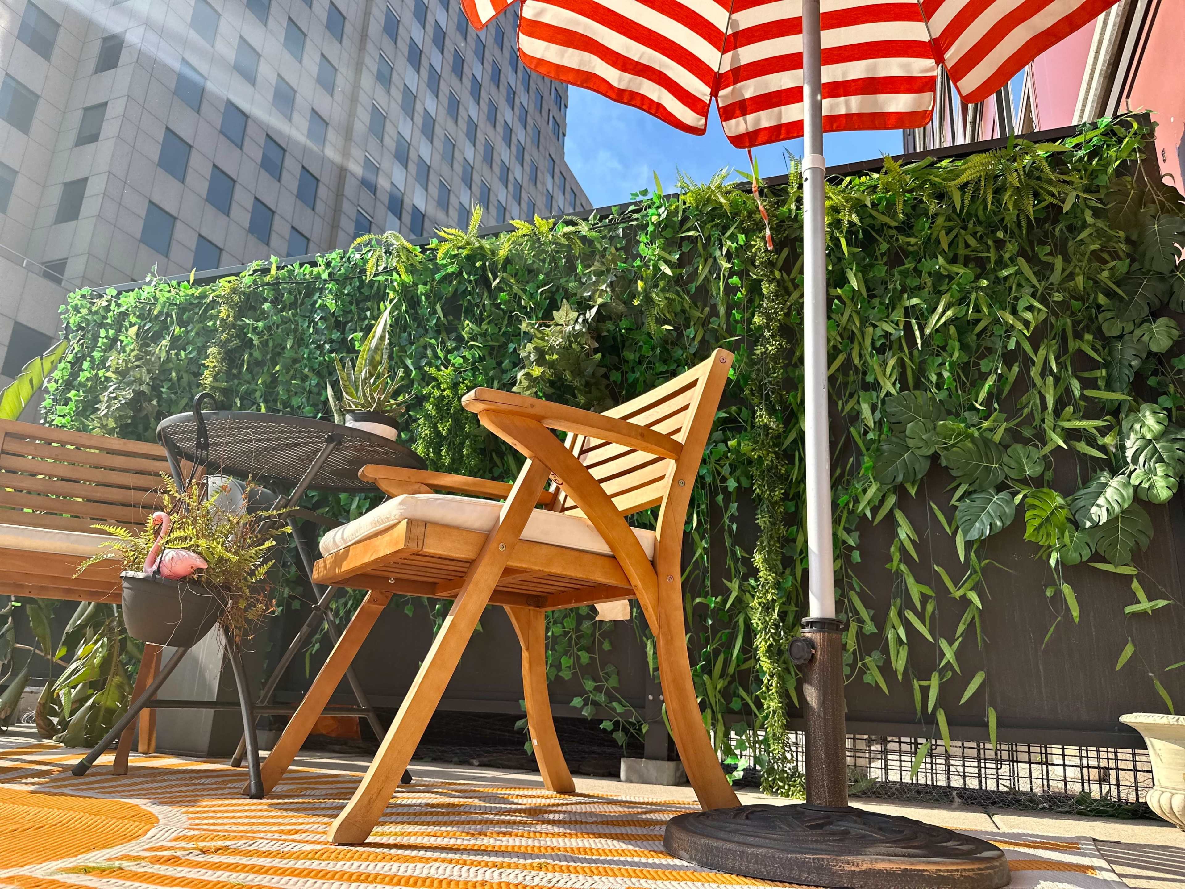 A wooden chair and table sit on a patterned rug beneath a striped umbrella, surrounded by artificial greenery on a balcony.