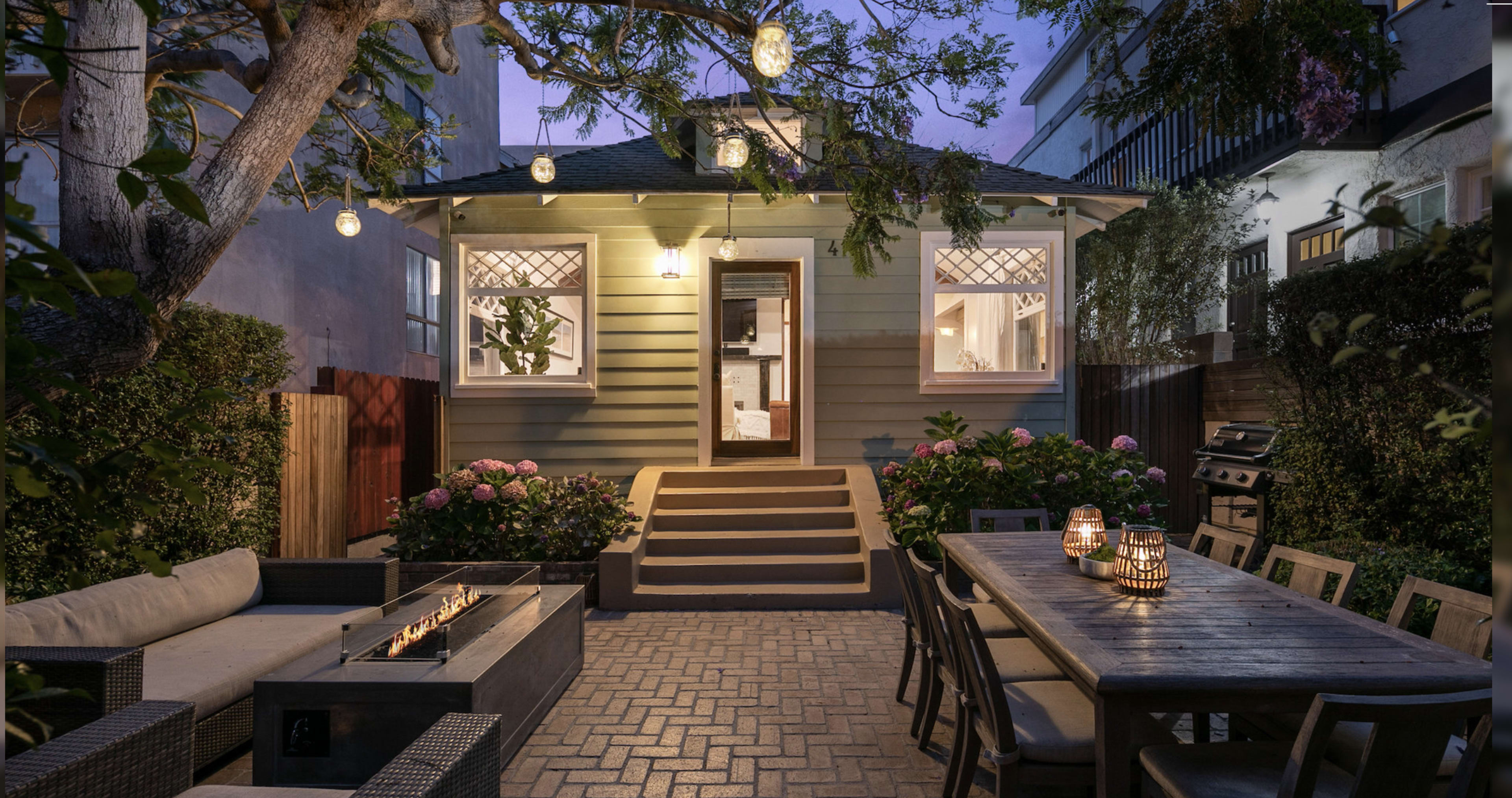 A cozy backyard features a seating area with a fire pit, a dining table, and string lights illuminating a charming home at dusk.