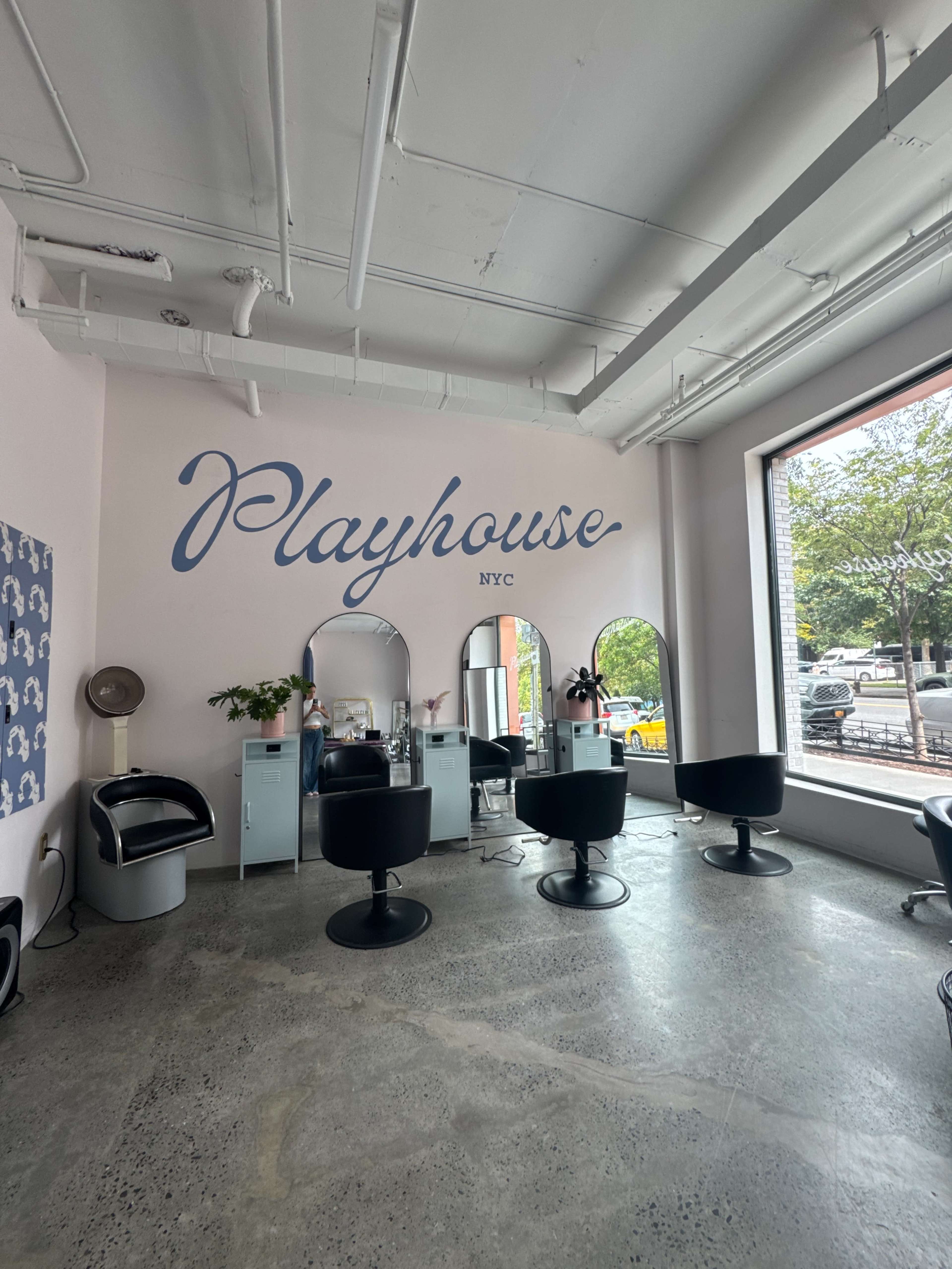 The interior of a hair salon named "Playhouse NYC" features several styling stations with mirrors and black chairs, illuminated by natural light from large windows.