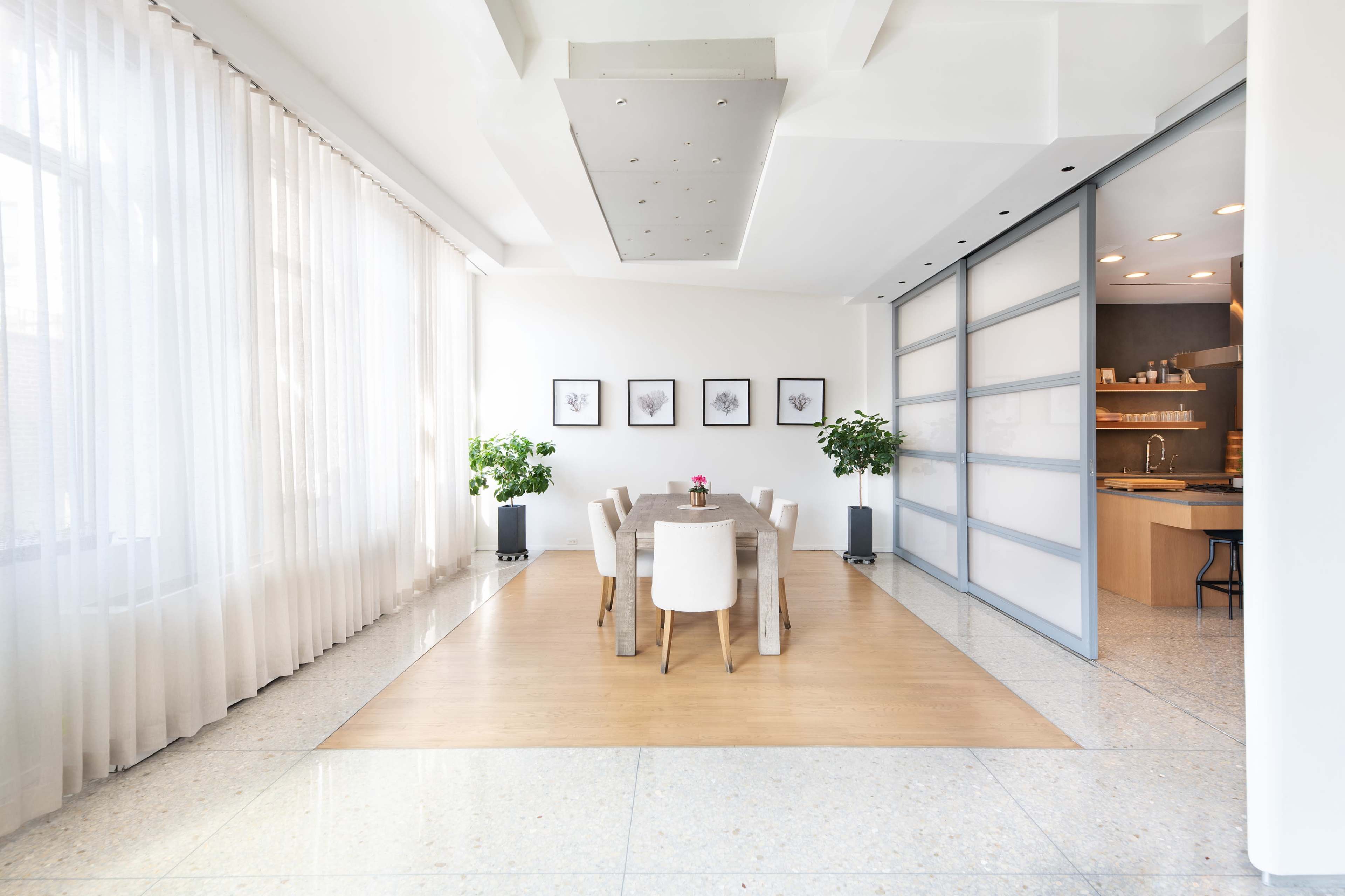 A dining area features a wooden table surrounded by white chairs, illuminated by natural light from large windows.