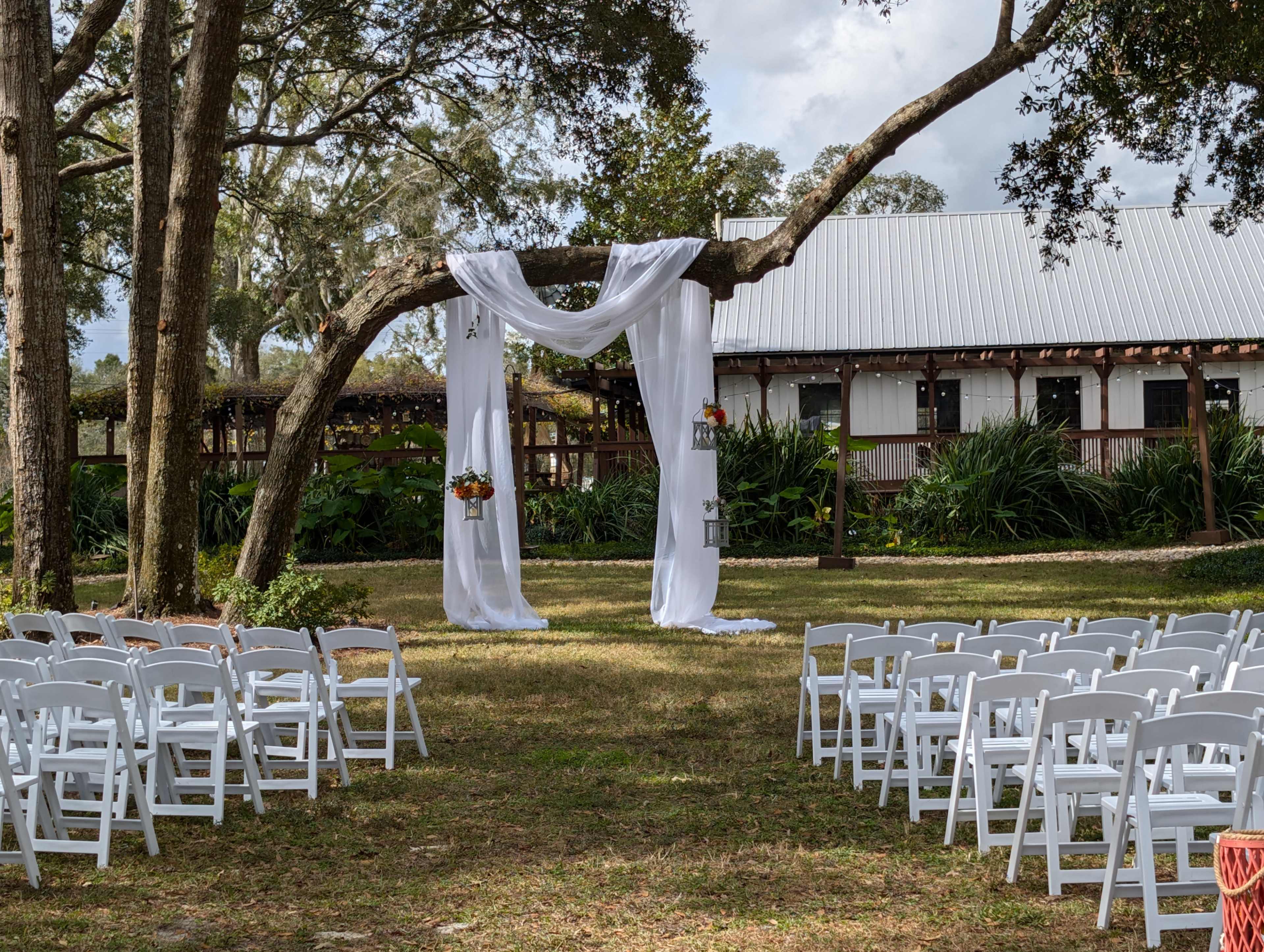A wedding ceremony setup features a white draped archway under a tree, surrounded by rows of white chairs on a grassy area.