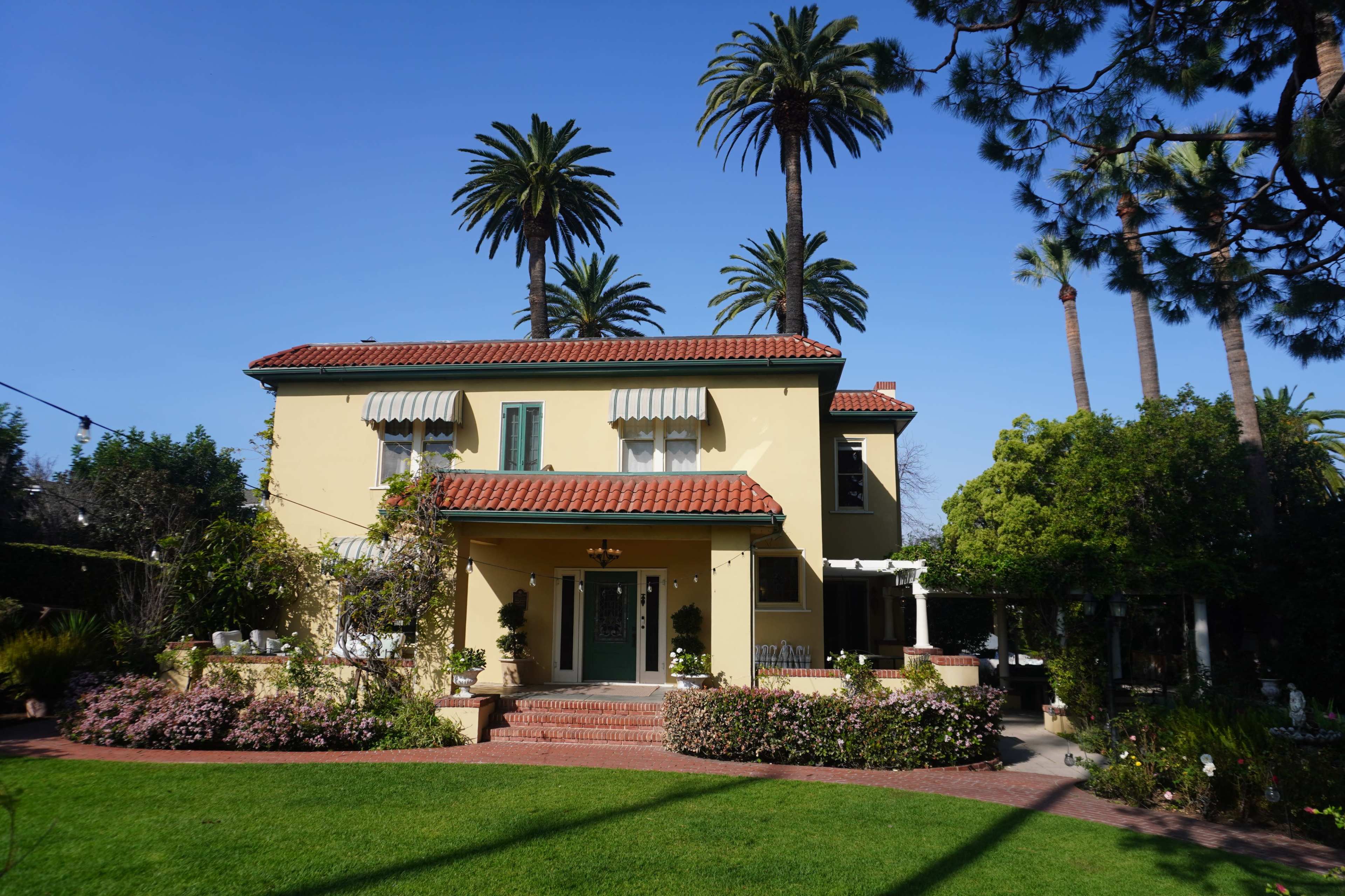 A two-story house with a yellow exterior and red-tiled roof is surrounded by tall palm trees and a landscaped garden.