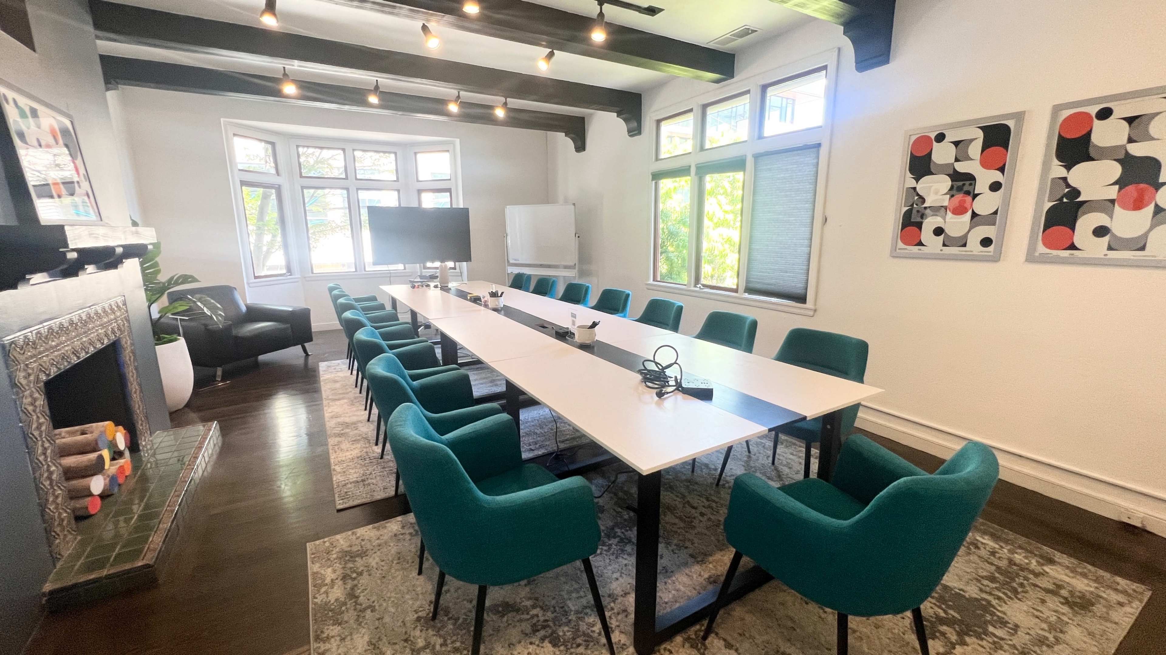A long conference table with teal chairs is set up in a room featuring a fireplace, large windows, and modern artwork on the walls.