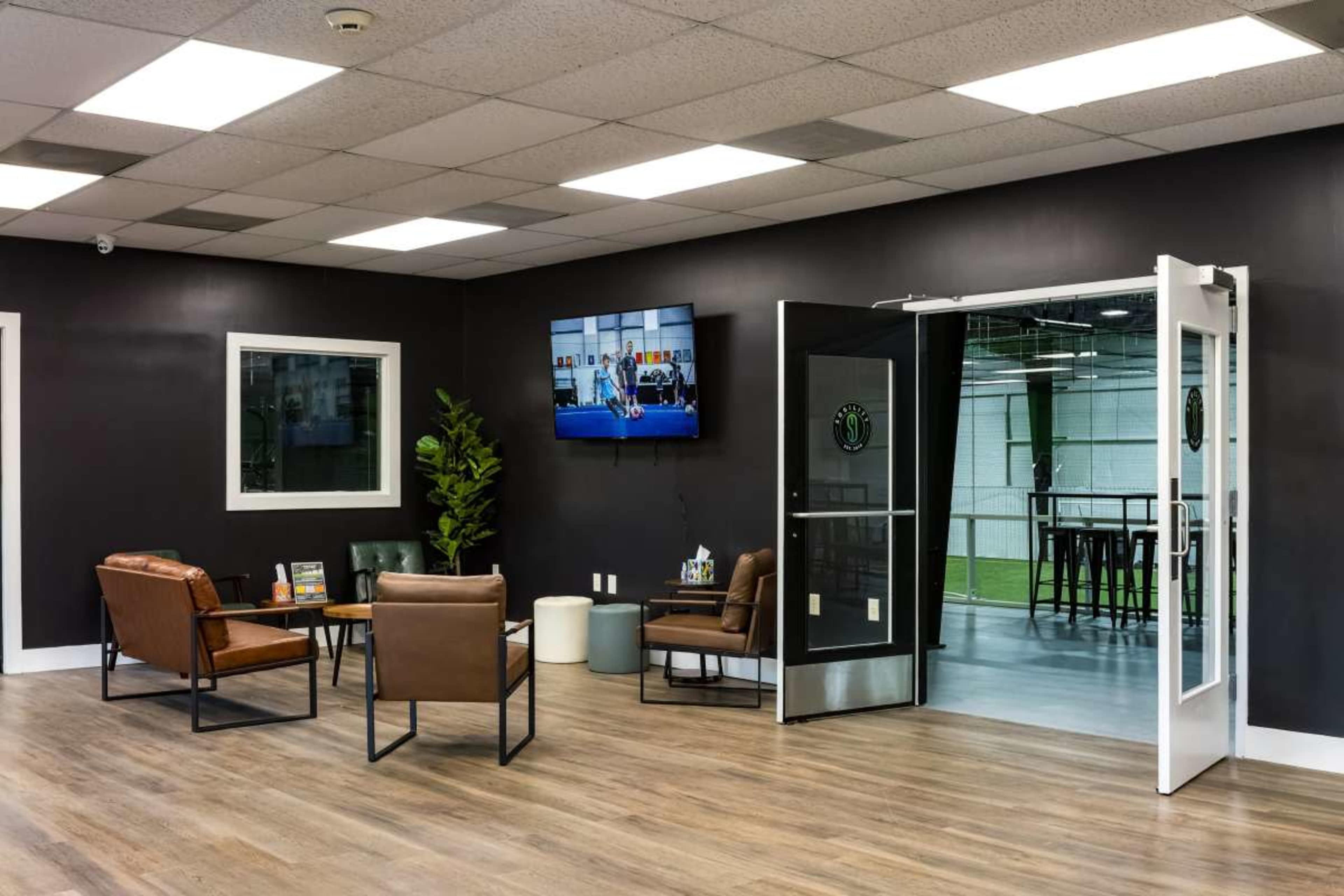The image shows a modern lounge area with leather chairs, a television mounted on the wall, and an open entrance leading to a spacious indoor facility.