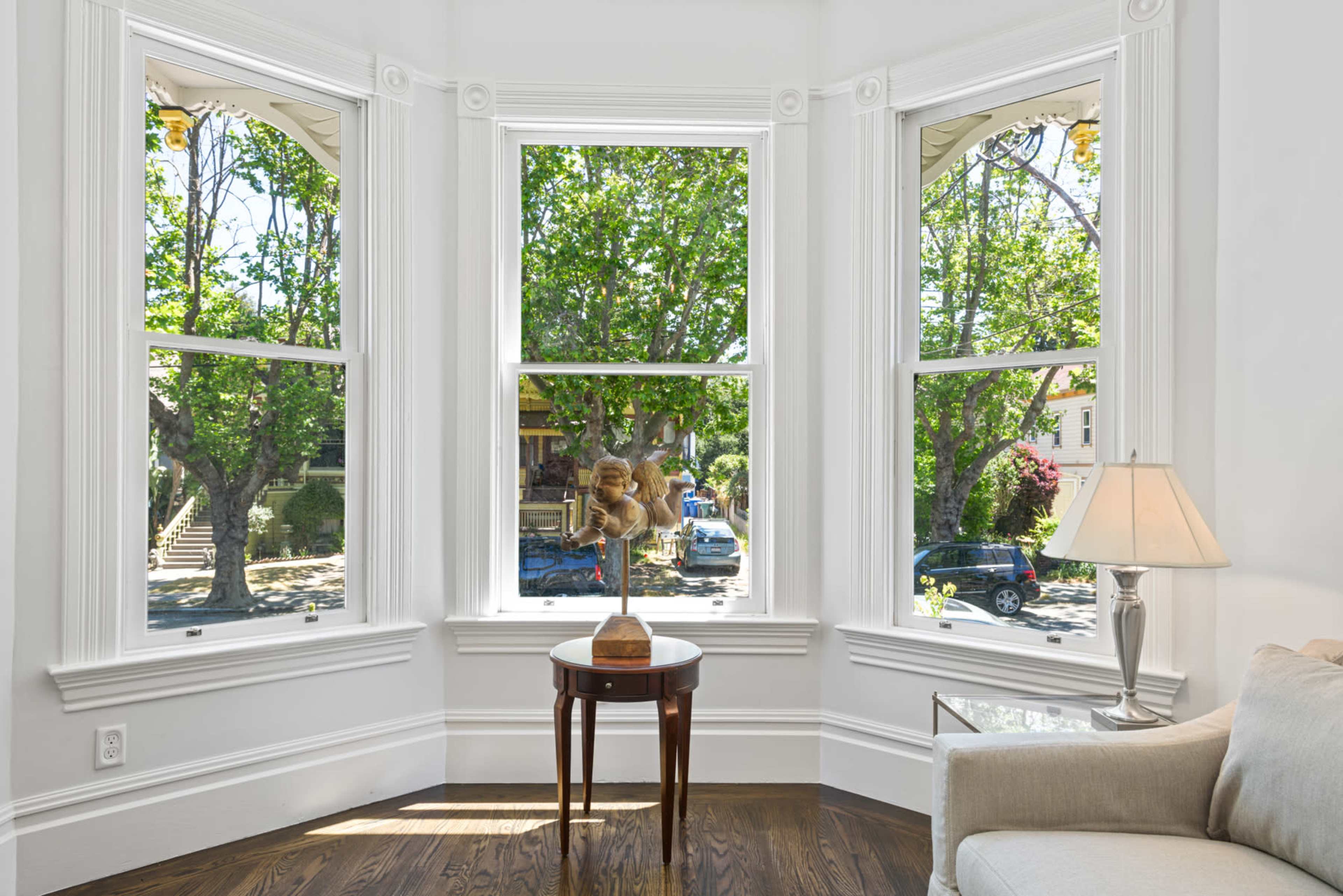 A small wooden table with a decorative object sits in front of three large windows that frame a view of trees and parked cars outside.