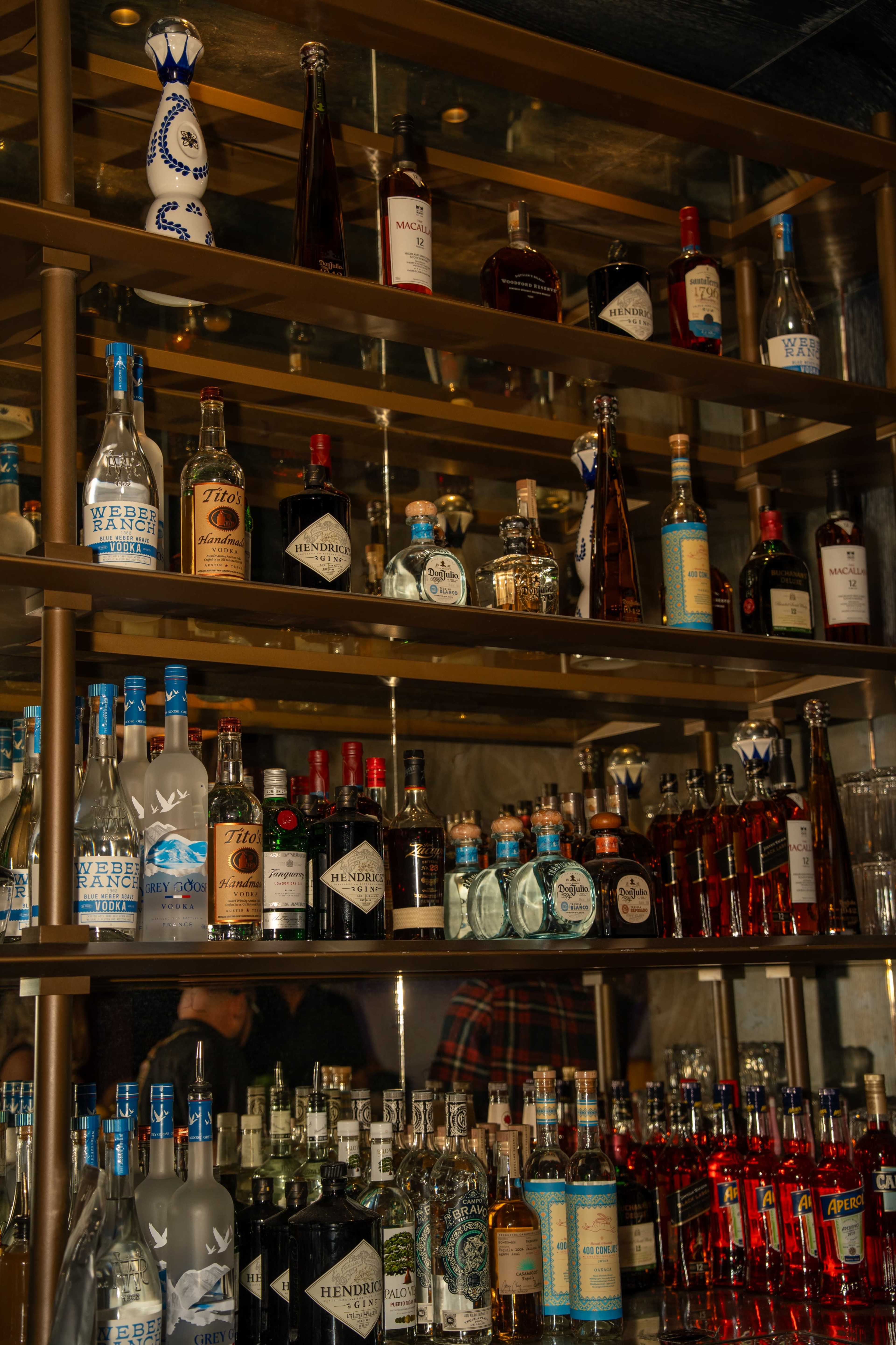 The image shows a neatly arranged shelf of various alcoholic beverages, including bottles of vodka, gin, rum, and whiskey, displayed in a bar setting.