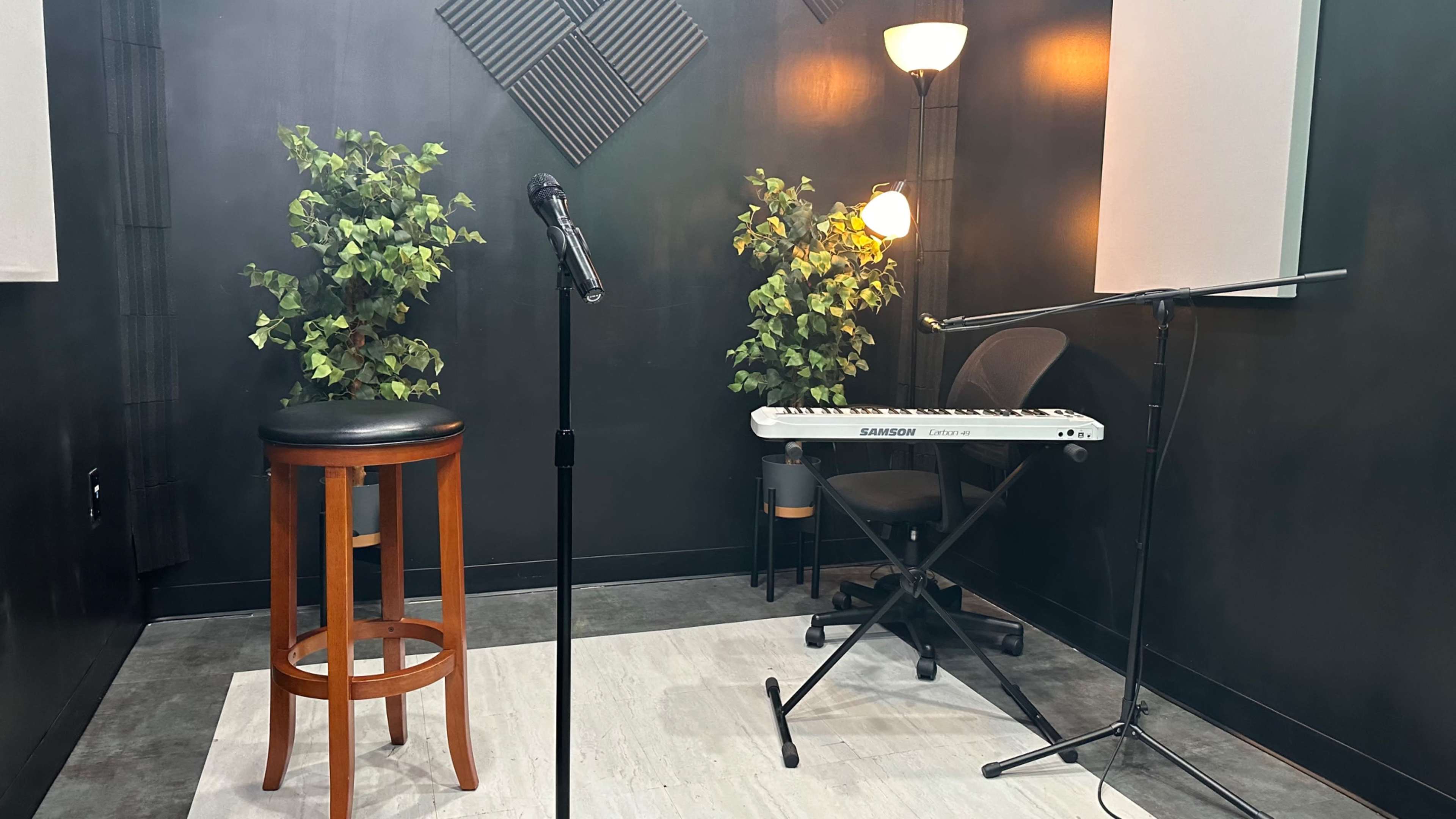 A small performance space with a microphone on a stand, a stool, a keyboard, and two potted plants against a dark wall.