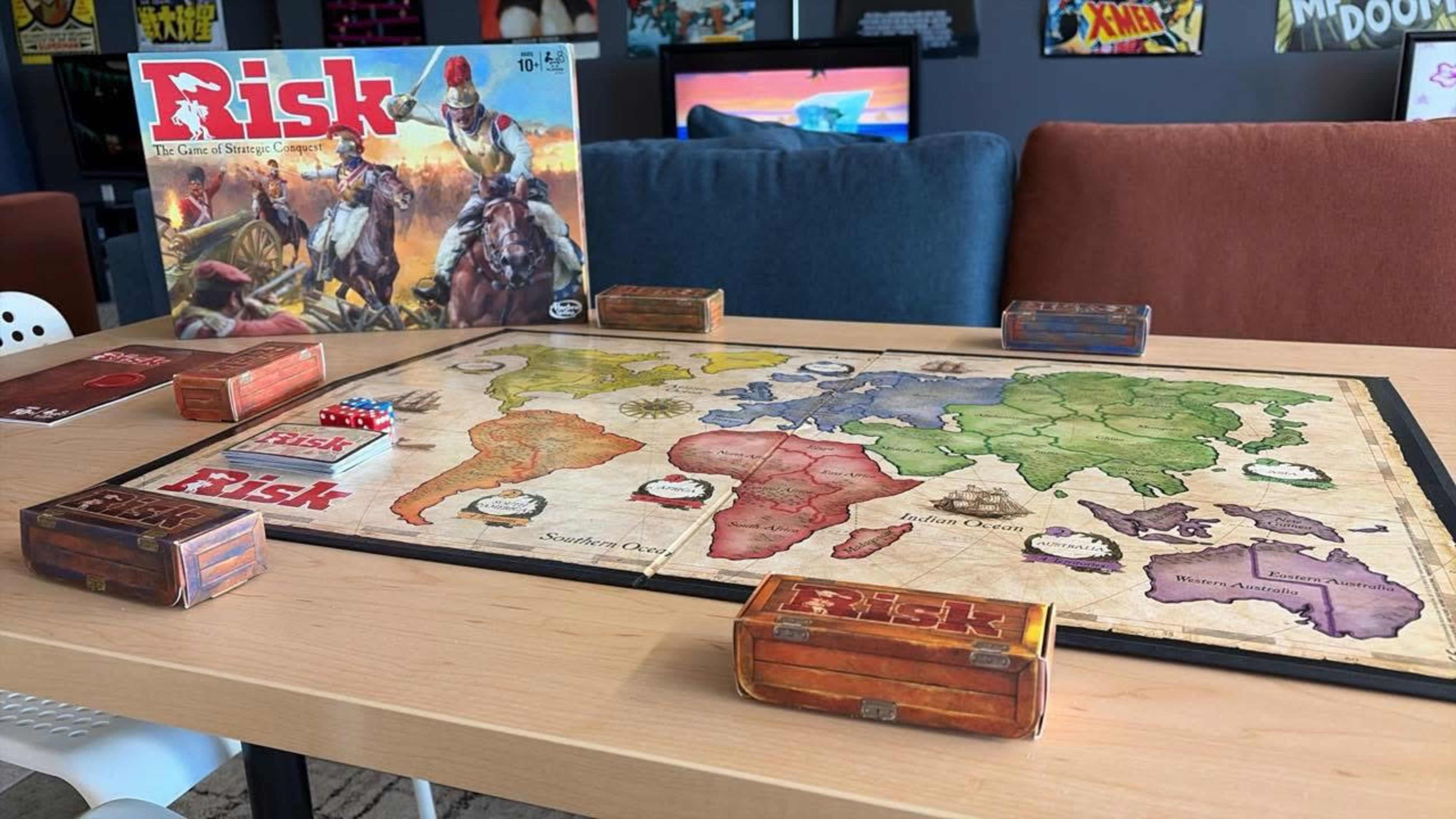 A Risk board game is set up on a table, featuring a world map and game pieces in wooden boxes, with the game box displayed in the background.